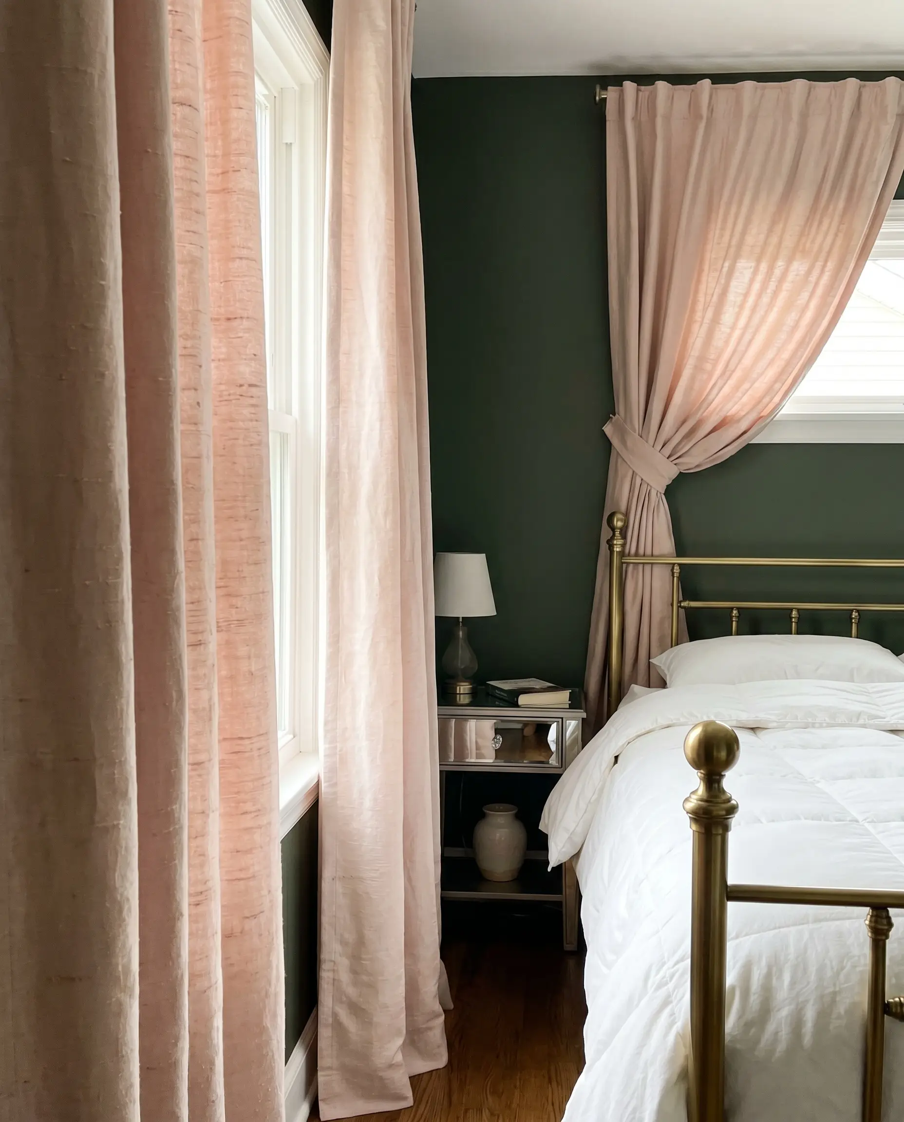 Heavyweight soft blush pink raw silk curtains frame a standard brass bed and mirrored nightstand against rich dark green bedroom walls.