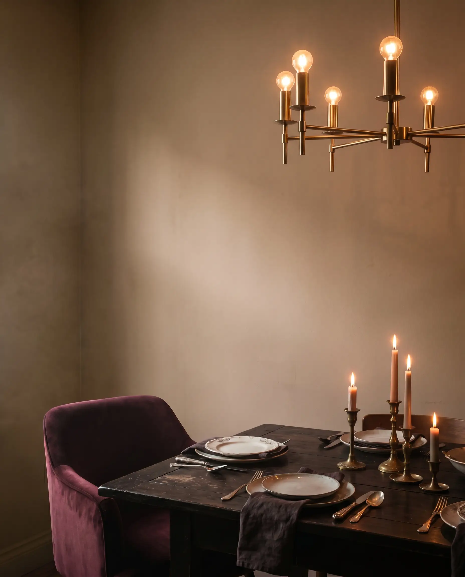 A warm brass chandelier and flickering taper candles illuminate a dark wood dining table and plush velvet chair surrounded by dusty pink-mushroom walls.