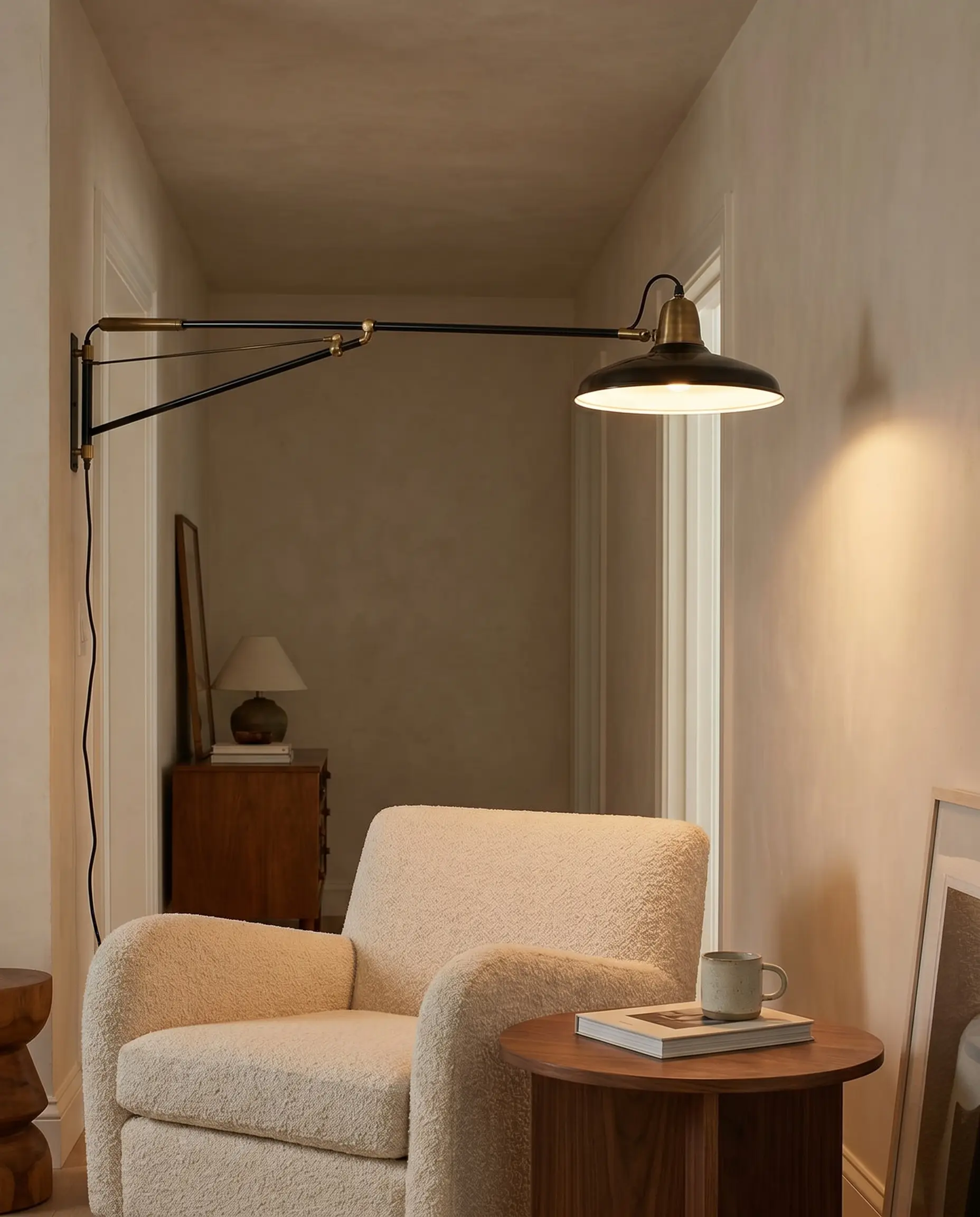 A double-jointed architectural wall sconce made of black enameled steel extends over a plush boucle lounge chair and small walnut side table in a reading nook.