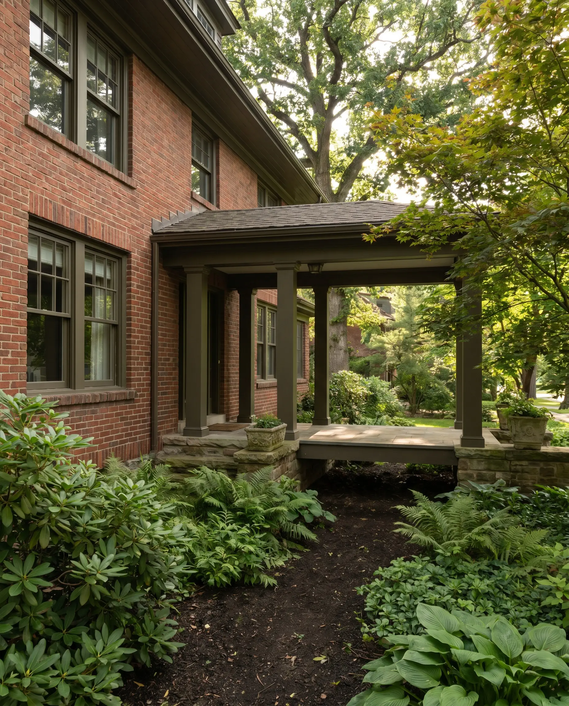 Dark olive-brown painted window trim and structural porch columns root a classic red brick home exterior into heavy mature foliage and shade plants.