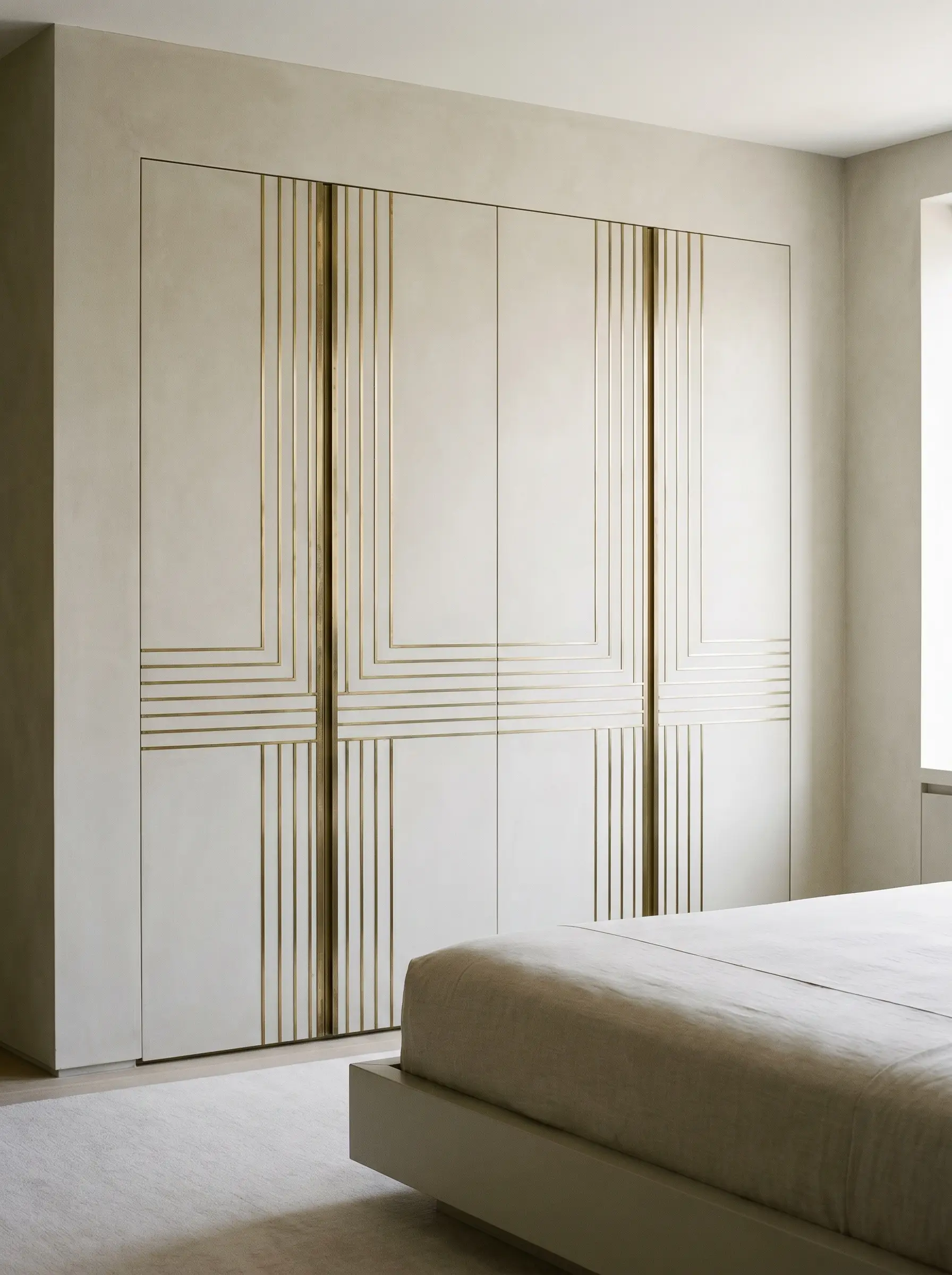 Flat-panel custom closet doors detailed with thin linear unlacquered brass inlays stand beside a neutral-toned wall and a sleek minimalist bed.