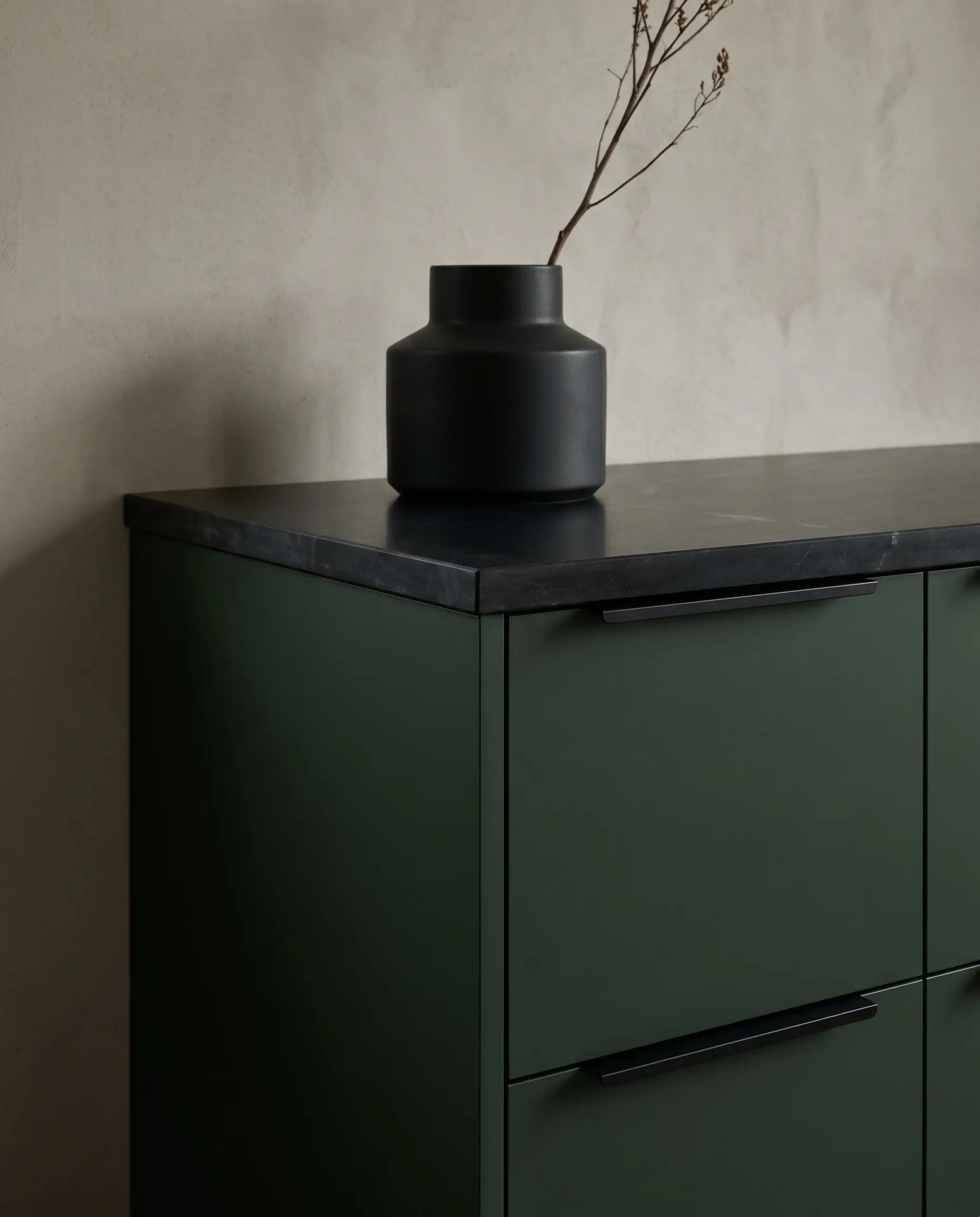 Flat-panel dark green cabinetry fitted with low-profile matte black edge pulls matches the square eased edge of an oiled black soapstone countertop holding a ceramic vase.