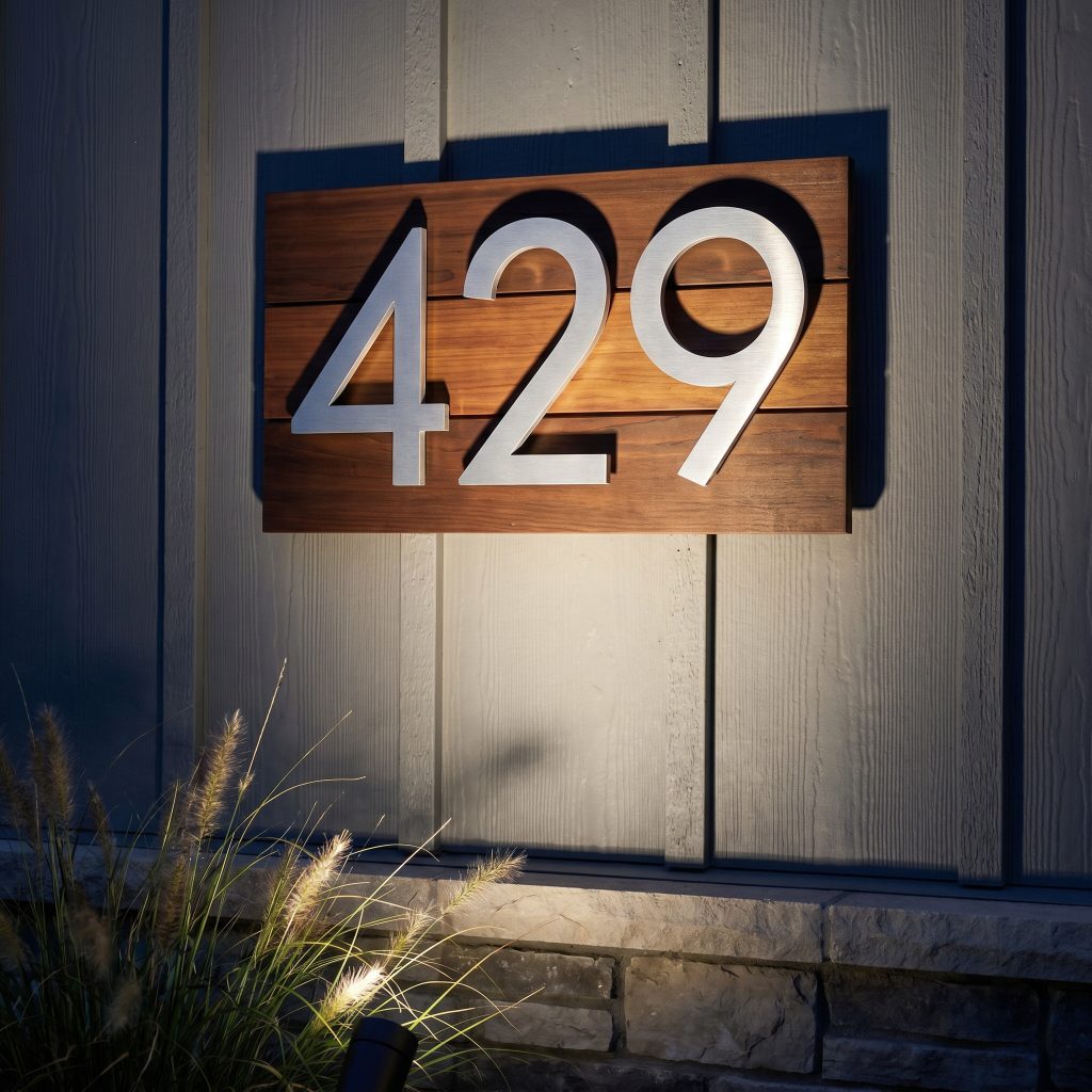 Oversized machined aluminum house numbers float off a warm cedar wood backplate mounted against textured board and batten siding, illuminated by a low-voltage spotlight.