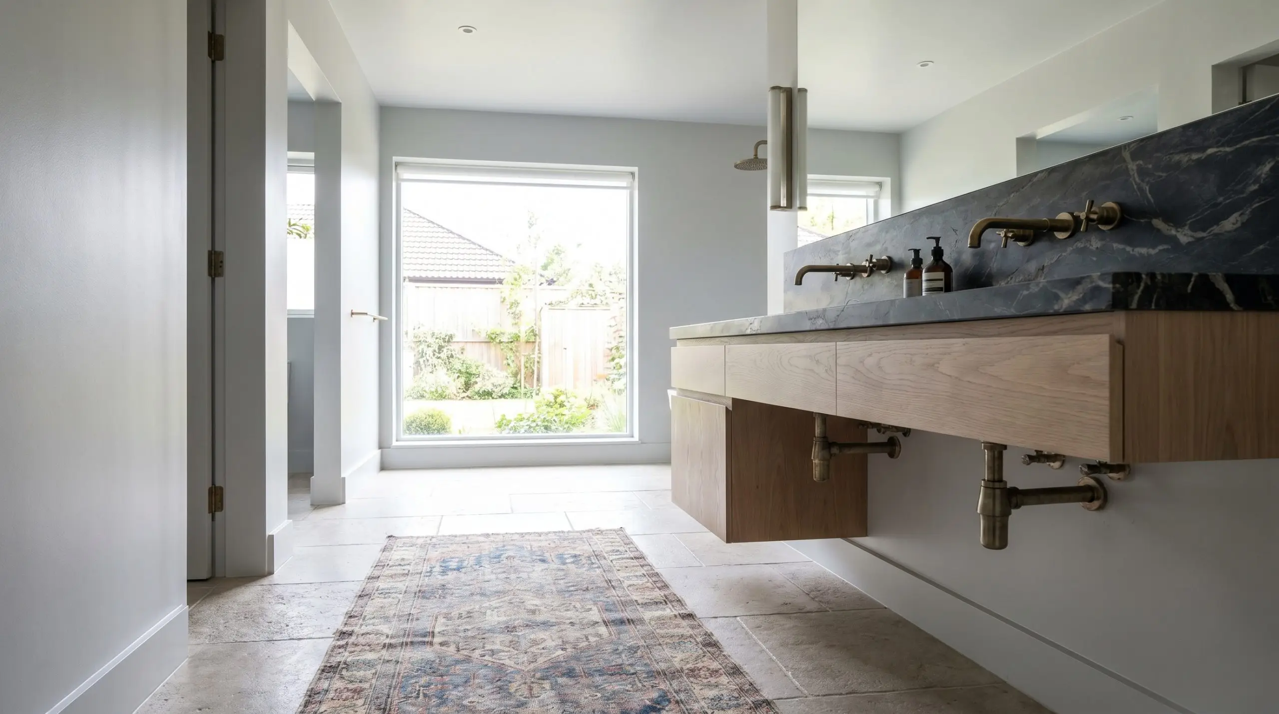 A floating white oak vanity with heavily veined soapstone counters sits inside a spacious wet room featuring cool gray walls and unlacquered brass plumbing fixtures.