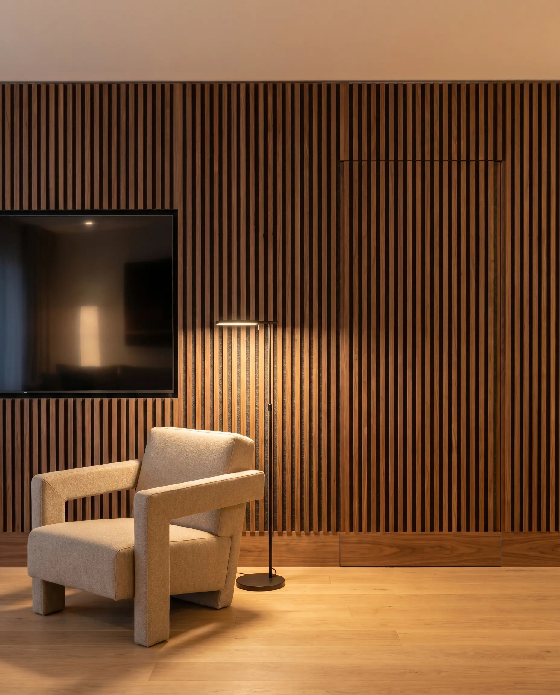 A flush-mount hidden door clad in vertical walnut battens blends seamlessly into a continuous acoustic slat wall beside a flat-screen television and structured armchair.