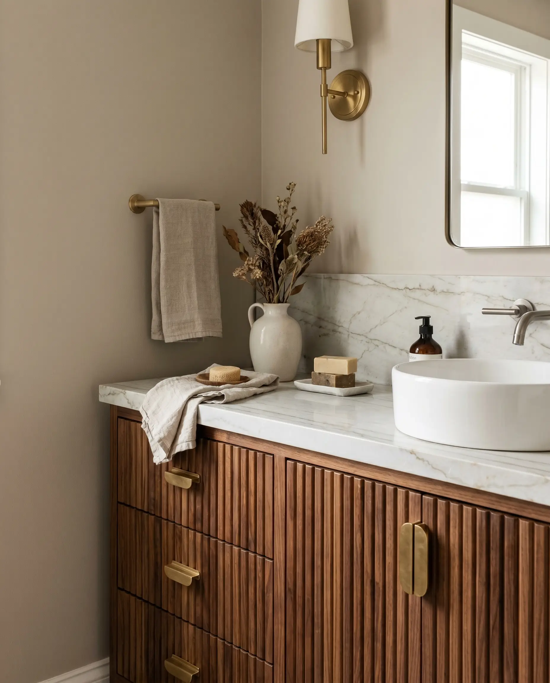 A solid walnut vanity with ribbed fluted detailing and unlacquered brass drawer pulls sits beneath a white ceramic vessel sink and marble countertop.