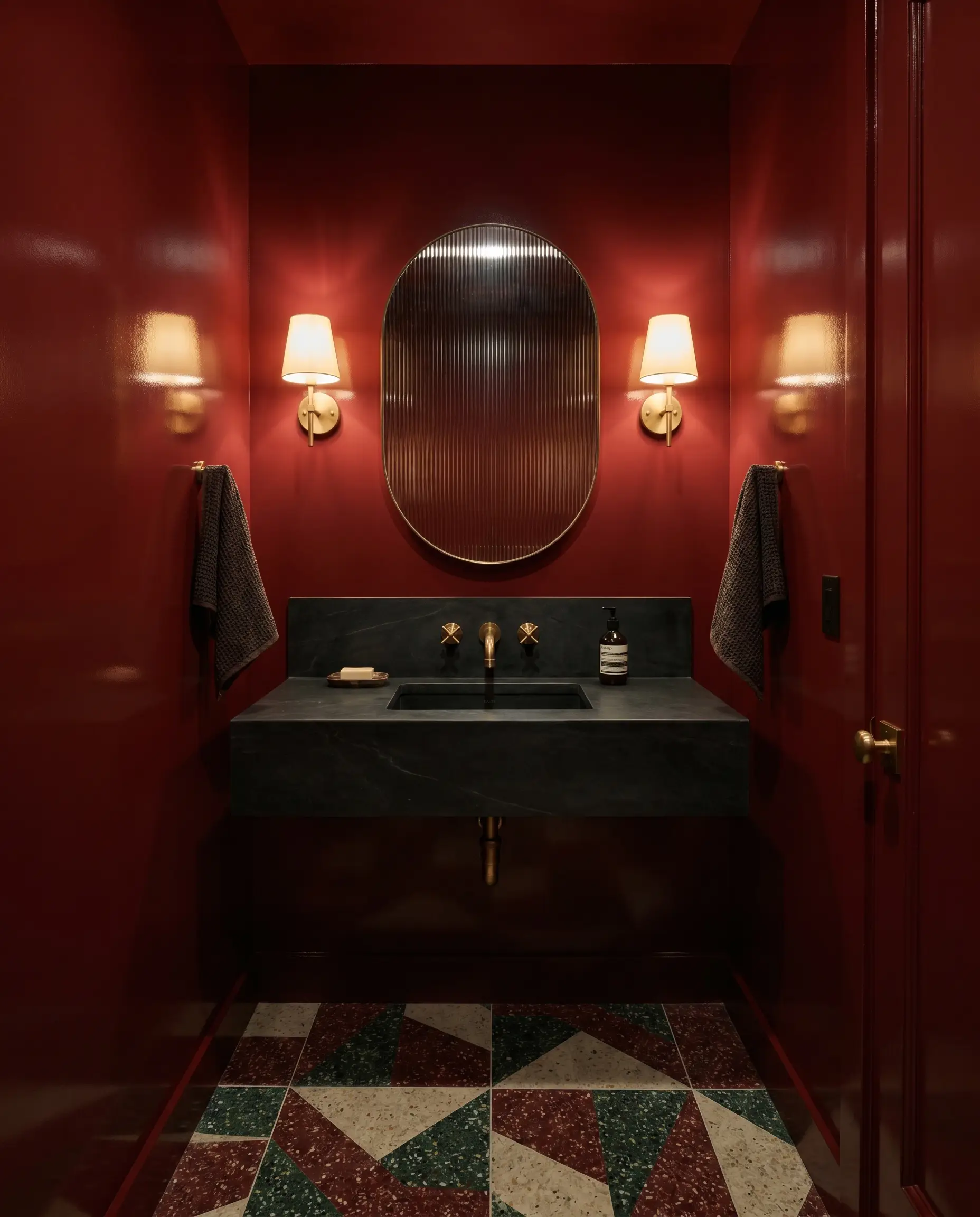 High-gloss lacquer crimson walls create a moody interior in a windowless powder room featuring a dark soapstone vanity, patterned terrazzo flooring, and a fluted glass mirror.