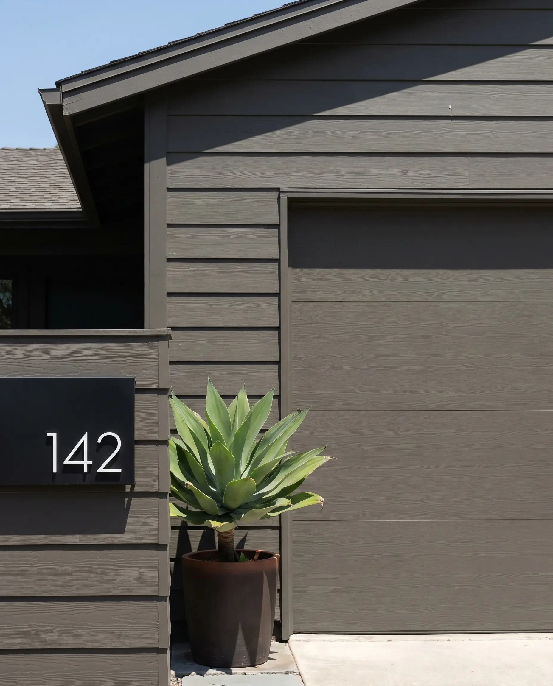 Modern greige facade ideas come to life on subtle lap siding featuring a modern matte black house number plaque and a sleek potted agave plant.
