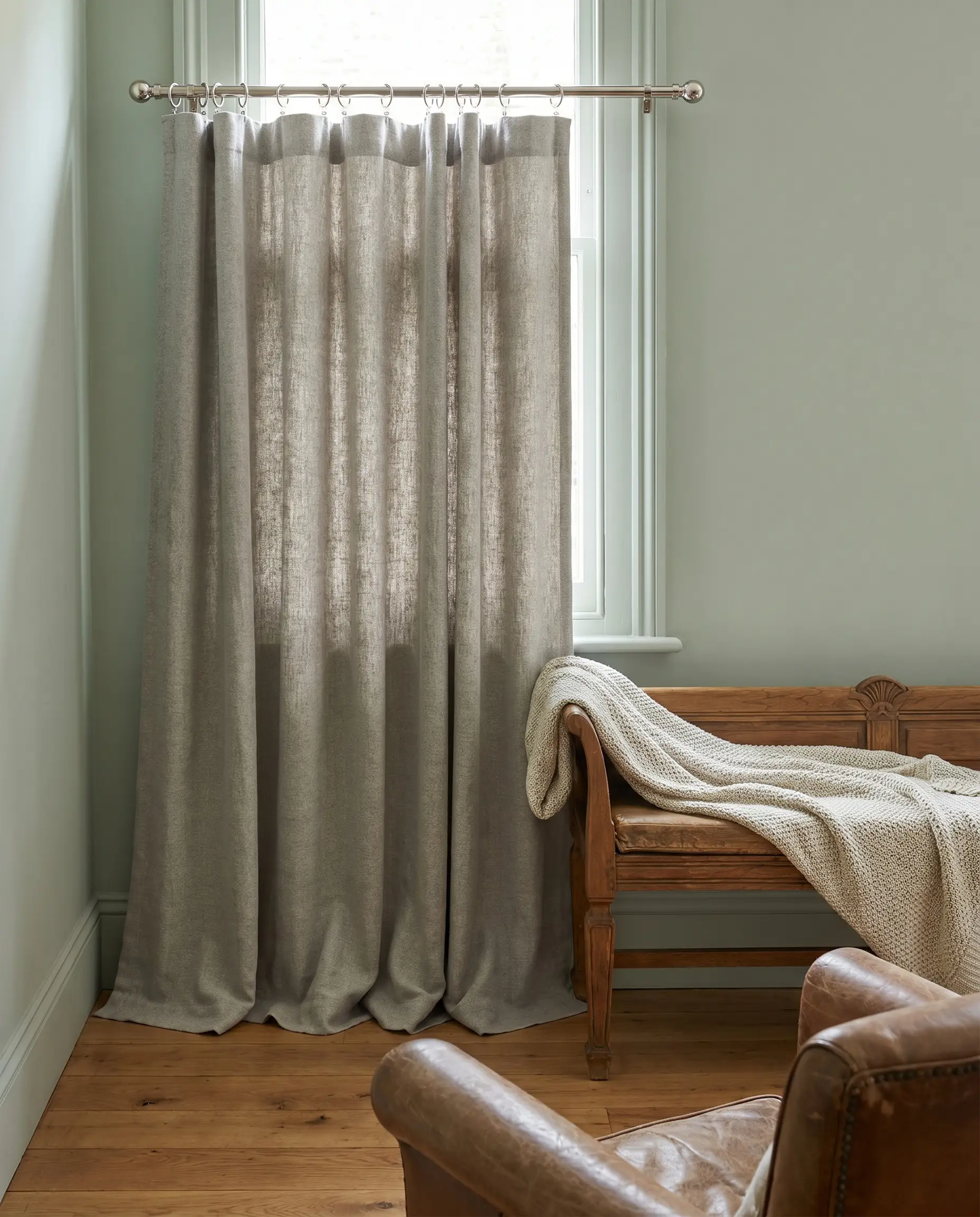 Heathered greige mid-weight linen curtains hang from a polished nickel rod with a puddle length break on warm oak floorboards near a vintage wooden bench.