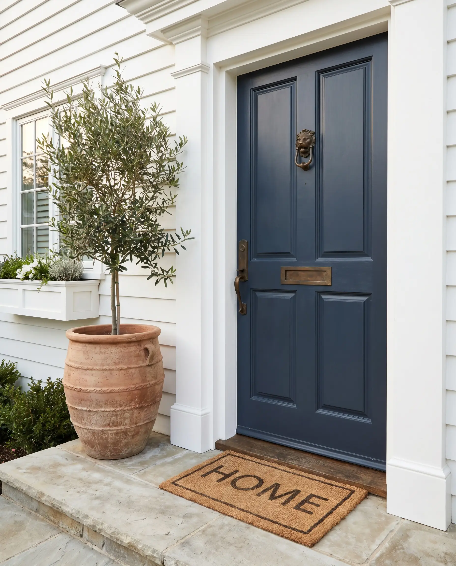 A deeply saturated coastal classic navy blue front door adorned with unlacquered brass hardware sits behind an inviting limestone porch step and textured ceramic planter.