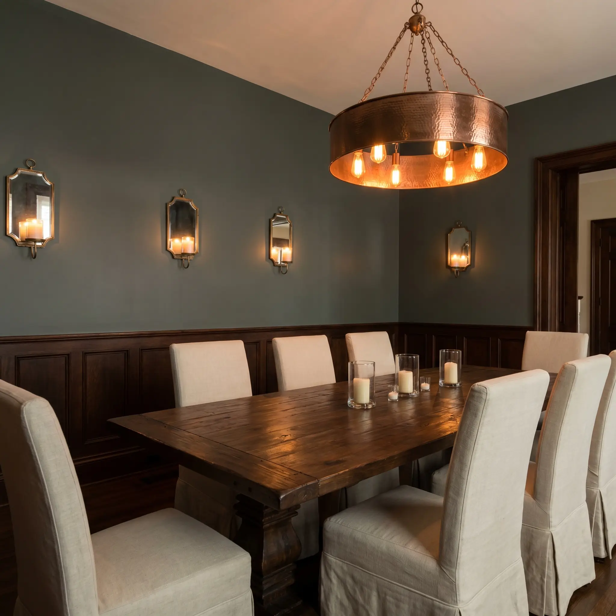 A hammered copper chandelier casts a warm glow over a dark walnut dining table and classic wainscoting painted in Farrow & Ball Douter (Hex #636f68).