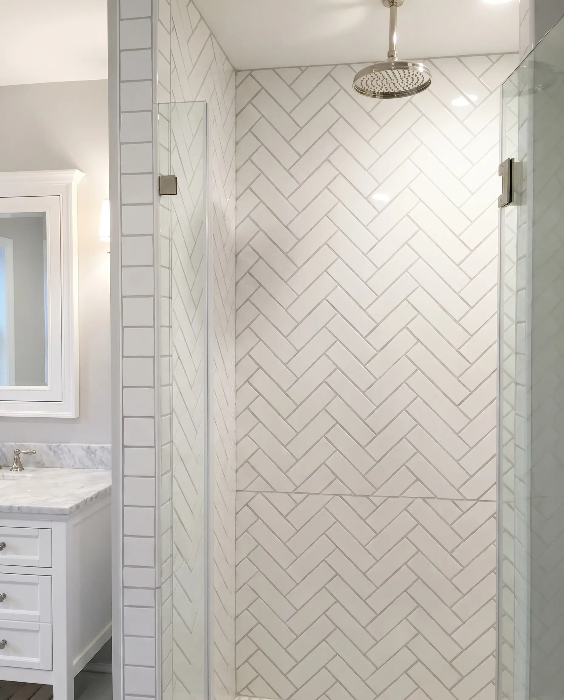 A spacious subway tile bathroom features a walk-in shower enclosure with a striking 45-degree herringbone pattern, light gray grout, and polished nickel rain showerhead.