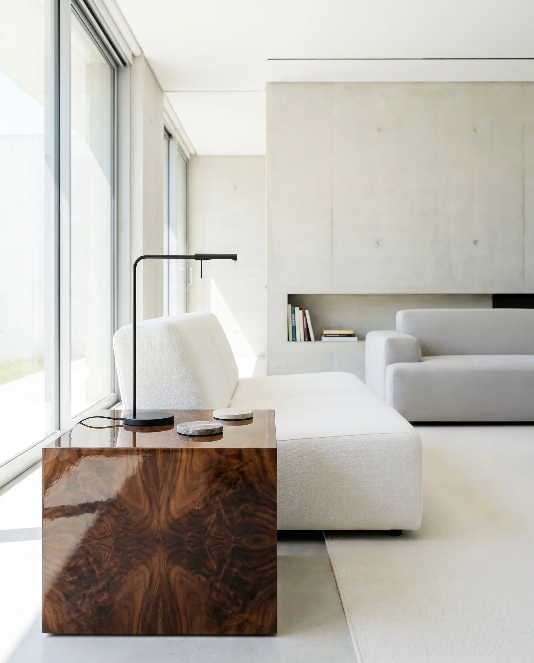 A high-gloss walnut burl wood side table holding a sleek contemporary reading lamp sits beside a minimalist white lounge chair.