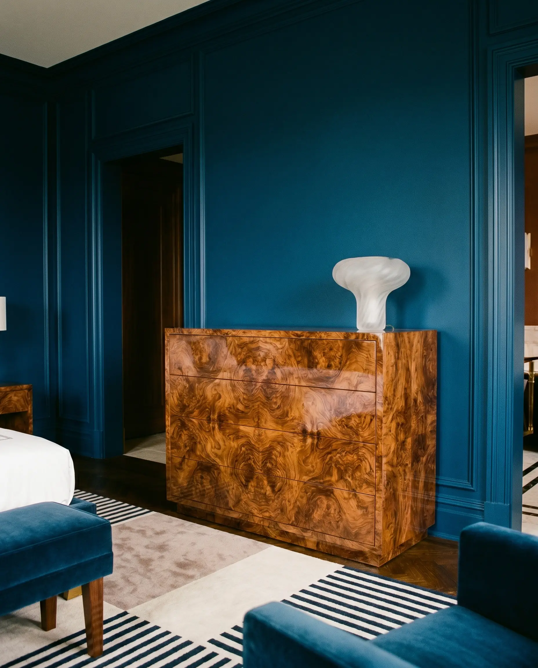 A large highly-figured burl wood veneer dresser stands as a statement piece against a solid jewel-toned wall, topped with a frosted glass lamp.
