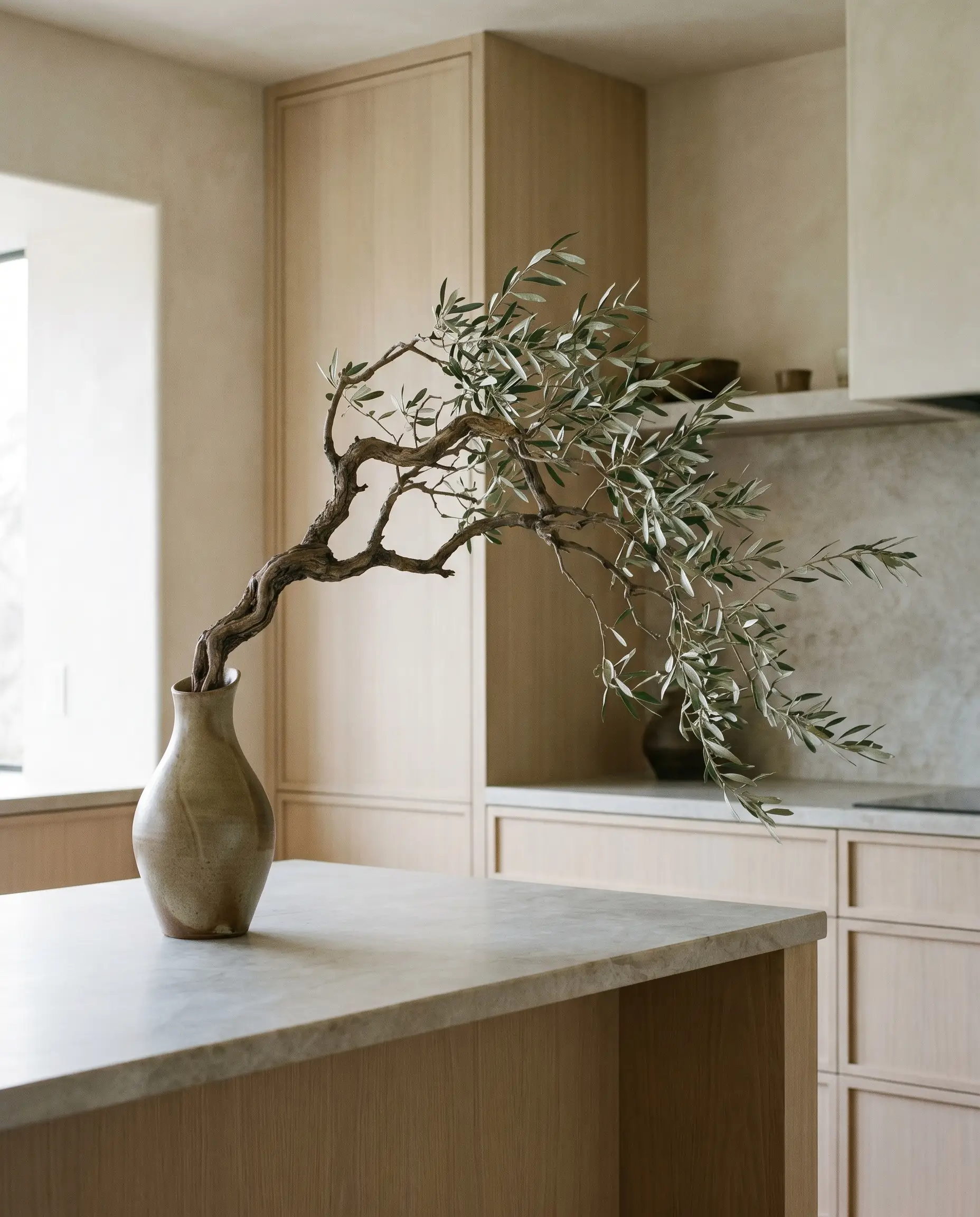 Japandi style kitchens embrace biophilic styling with a structural foraged olive branch in an asymmetrical ceramic vessel resting on a honed soapstone island.