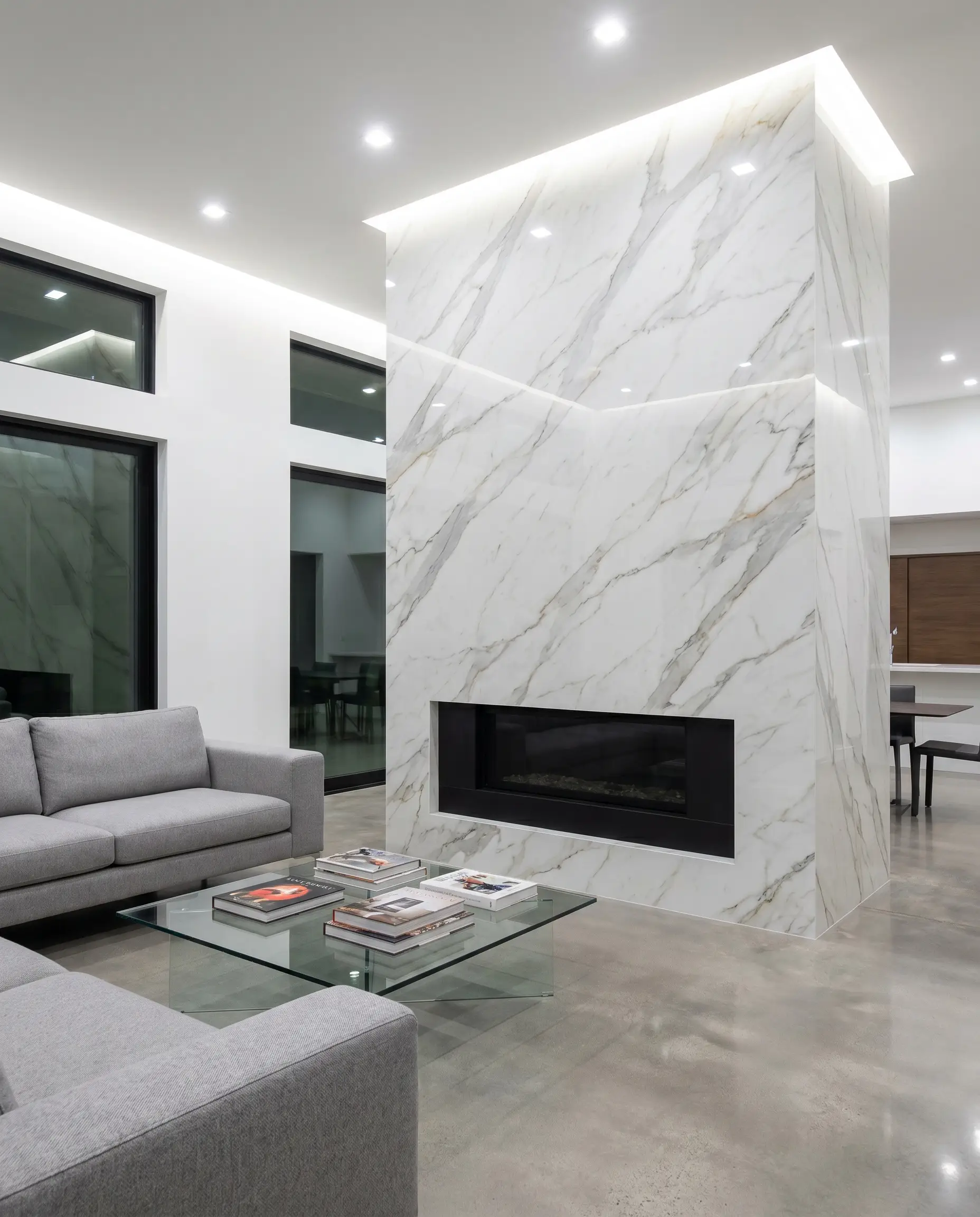 Ultra-compact large-format porcelain slabs with perfectly mitered edges mimic natural Statuario veining on a sleek firebox surround behind a minimalist grey sectional.