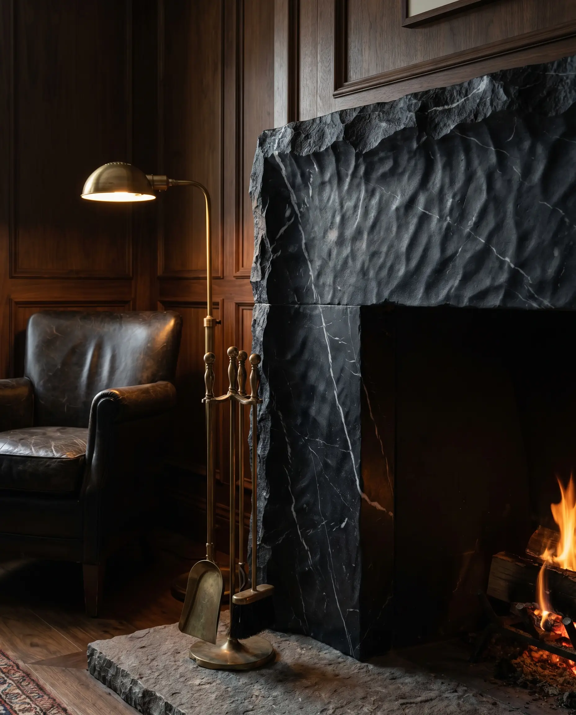 A deeply textured leathered Nero Marquina black stone surround stands against rich walnut paneling, accented by unlacquered brass tools and a dark leather club chair.