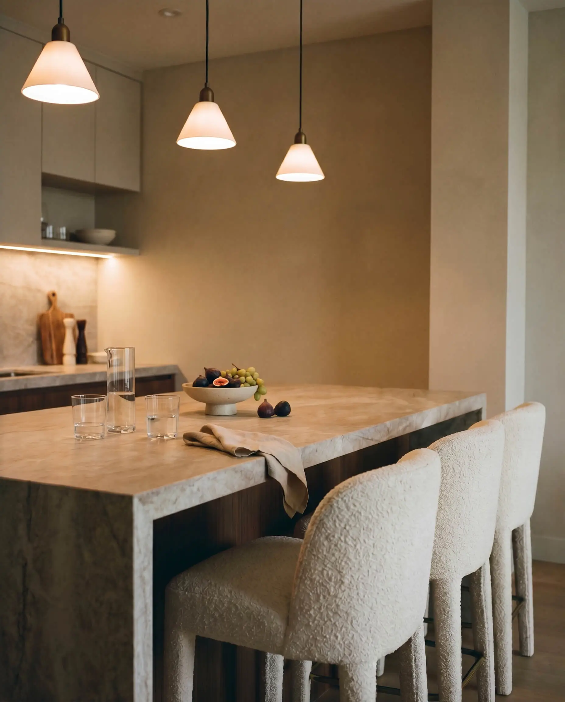 Plush upholstered barstools tuck beneath the cantilevered overhang of tactile Taj Mahal quartzite kitchen islands styled with modern glassware.