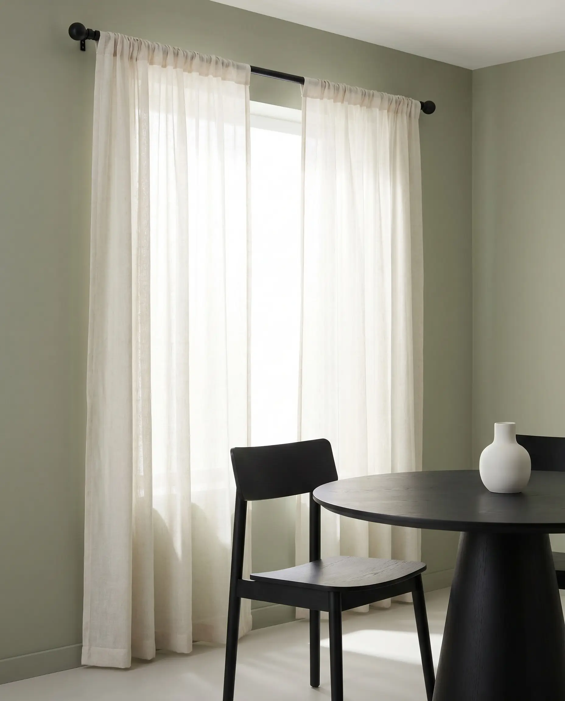 Alabaster light-filtering sheers hang from a matte black French return rod against painted green walls near a black wooden dining chair and pedestal table.