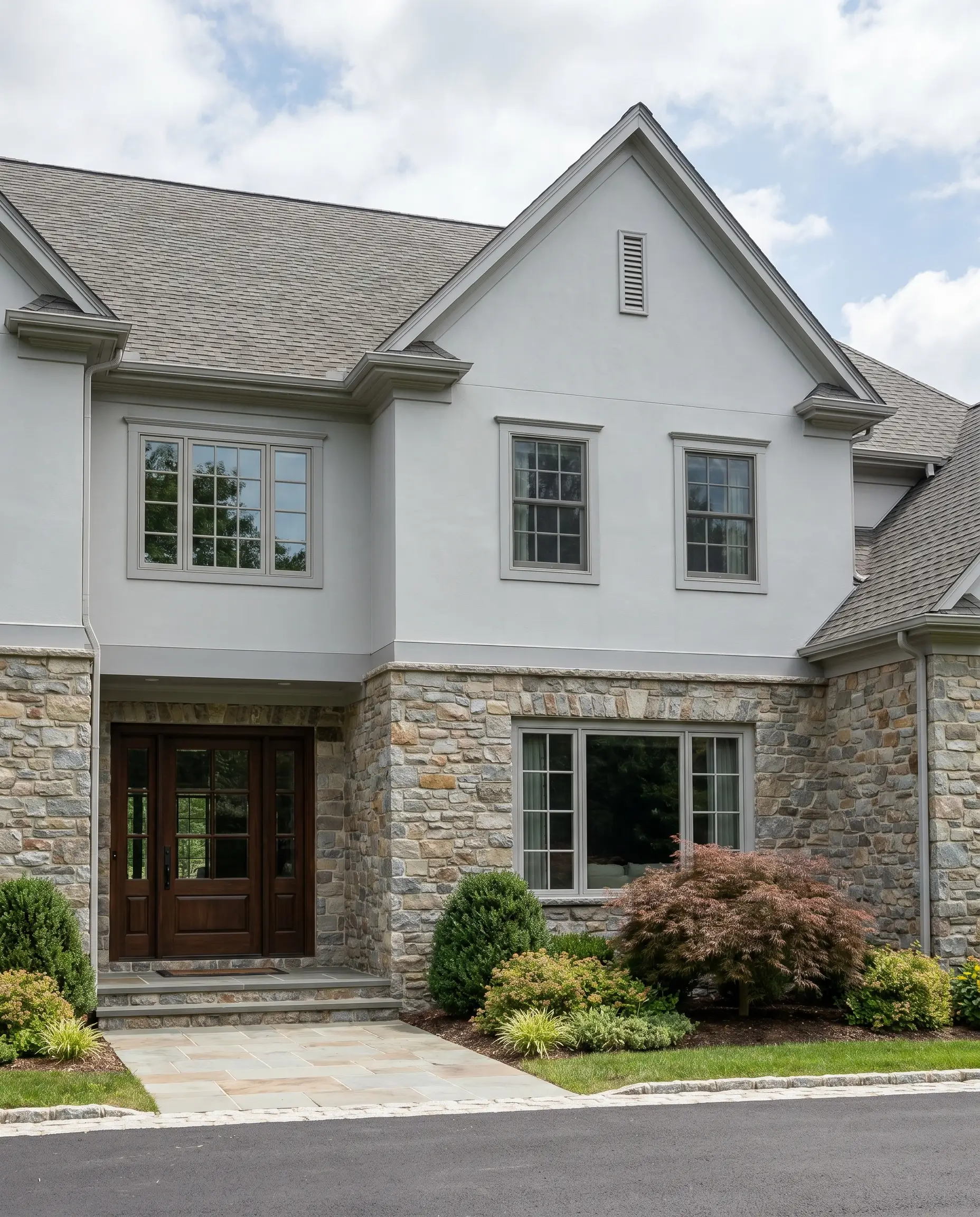 Smooth light gray siding clads the upper elevation of a stately home, resting above a heavy, dry-stacked fieldstone veneer base.