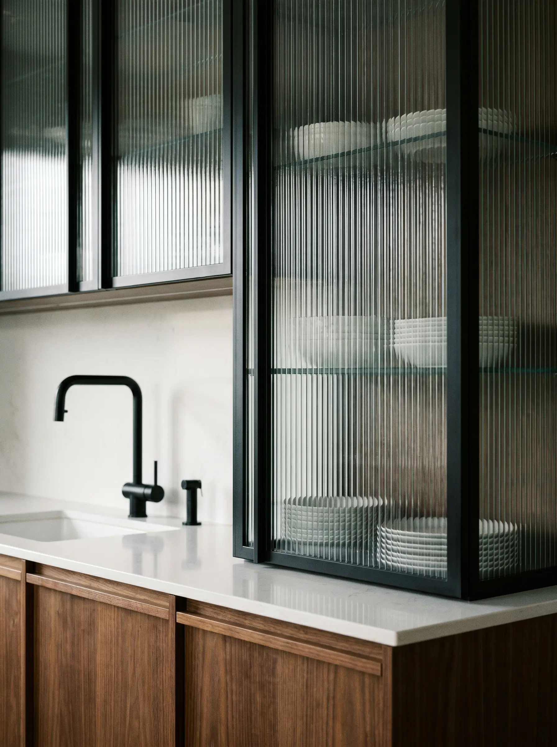 Slim matte black aluminum extrusion frames house ribbed glass upper cabinets above a white quartz countertop and rift-sawn wood base units.