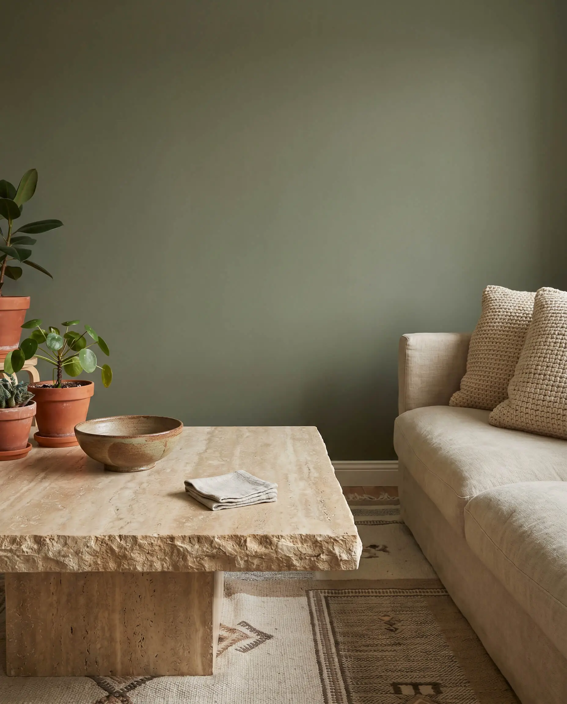 A honed matte travertine coffee table holding a ceramic bowl and linen coaster sits before a neutral linen sofa and flat earthy green wall.