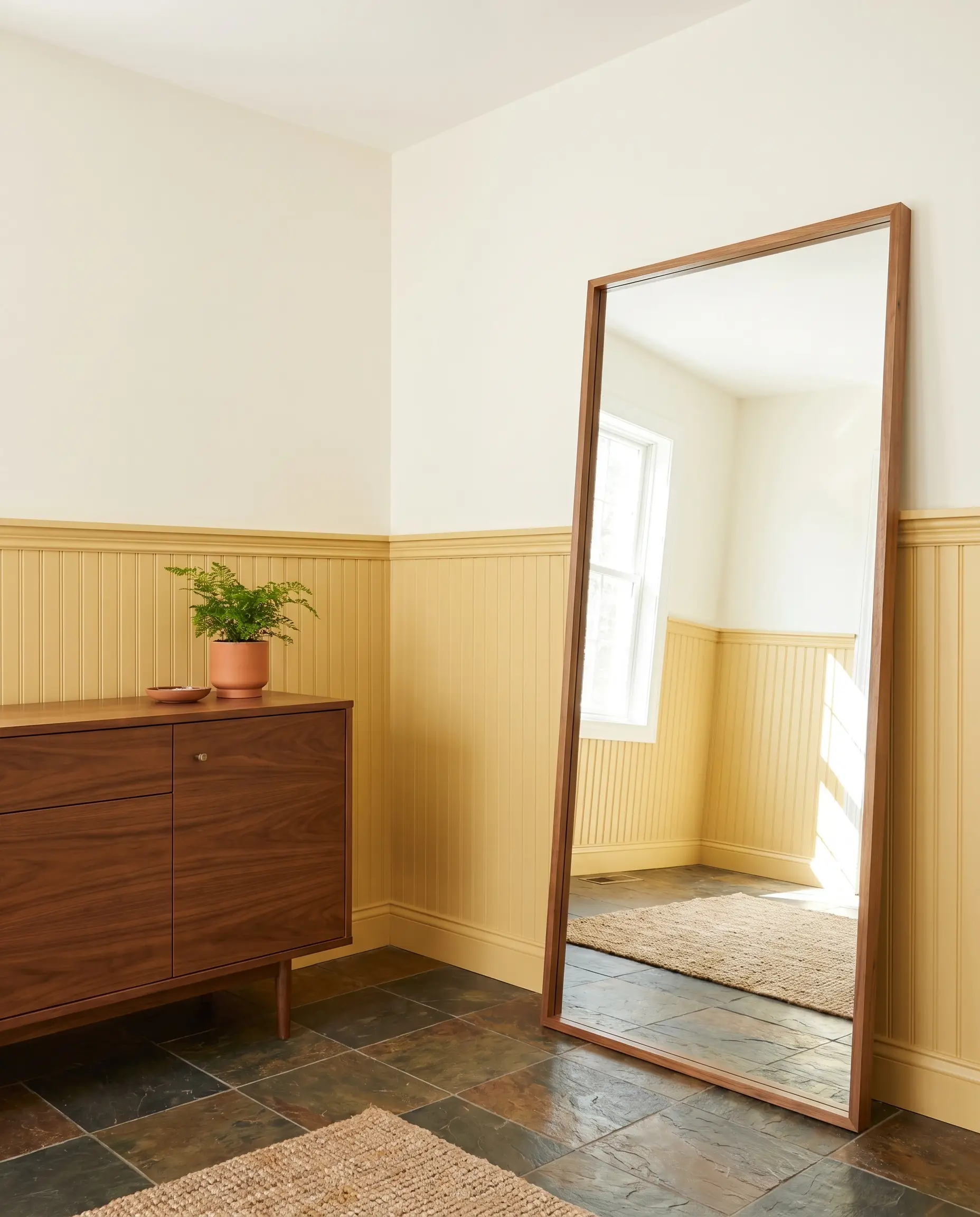 A sleek mid-century wooden credenza and oversized leaning floor mirror rest against lower beadboard wainscoting painted a warm historical ochre above earthy slate tiles.