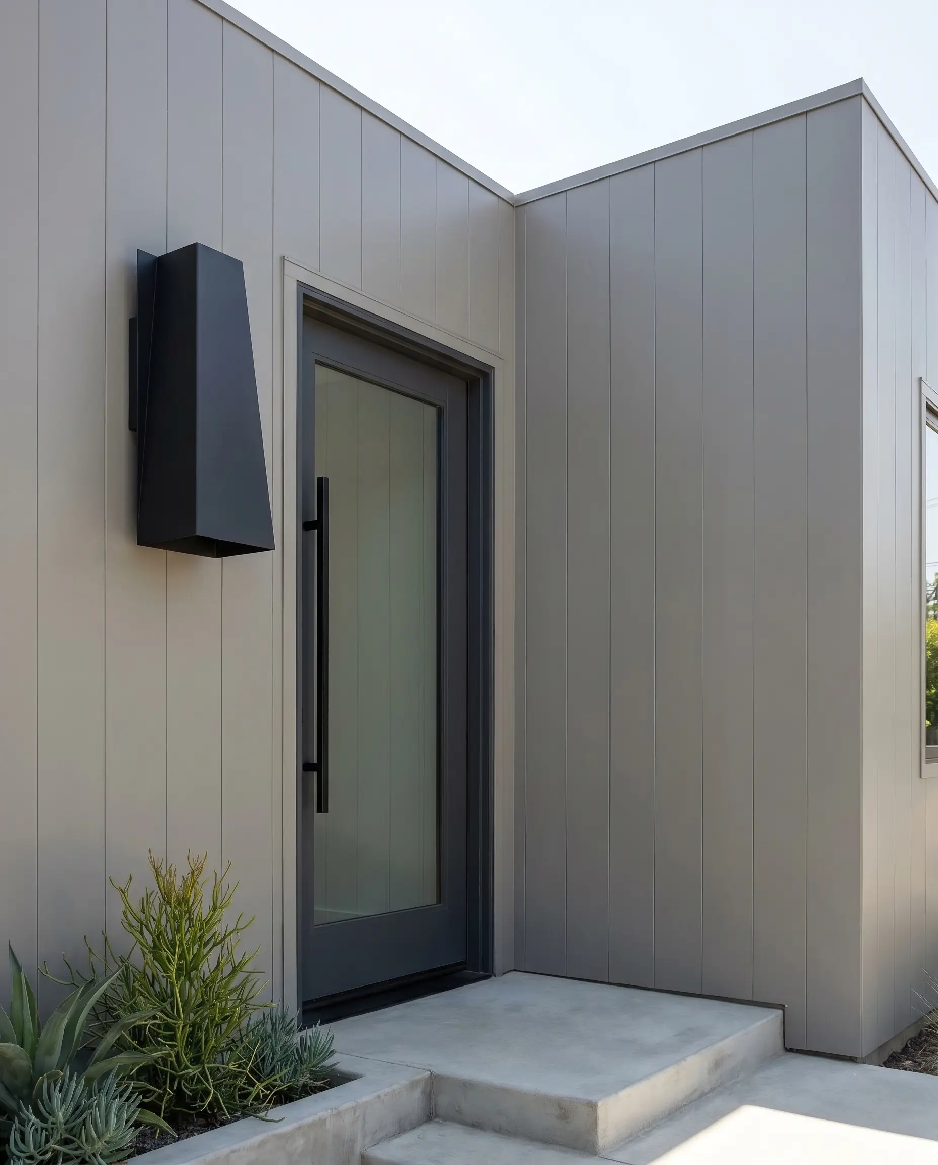An oversized matte black geometric sconce is proudly mounted beside a sleek door frame against crisp, mid-tone gray exterior siding.