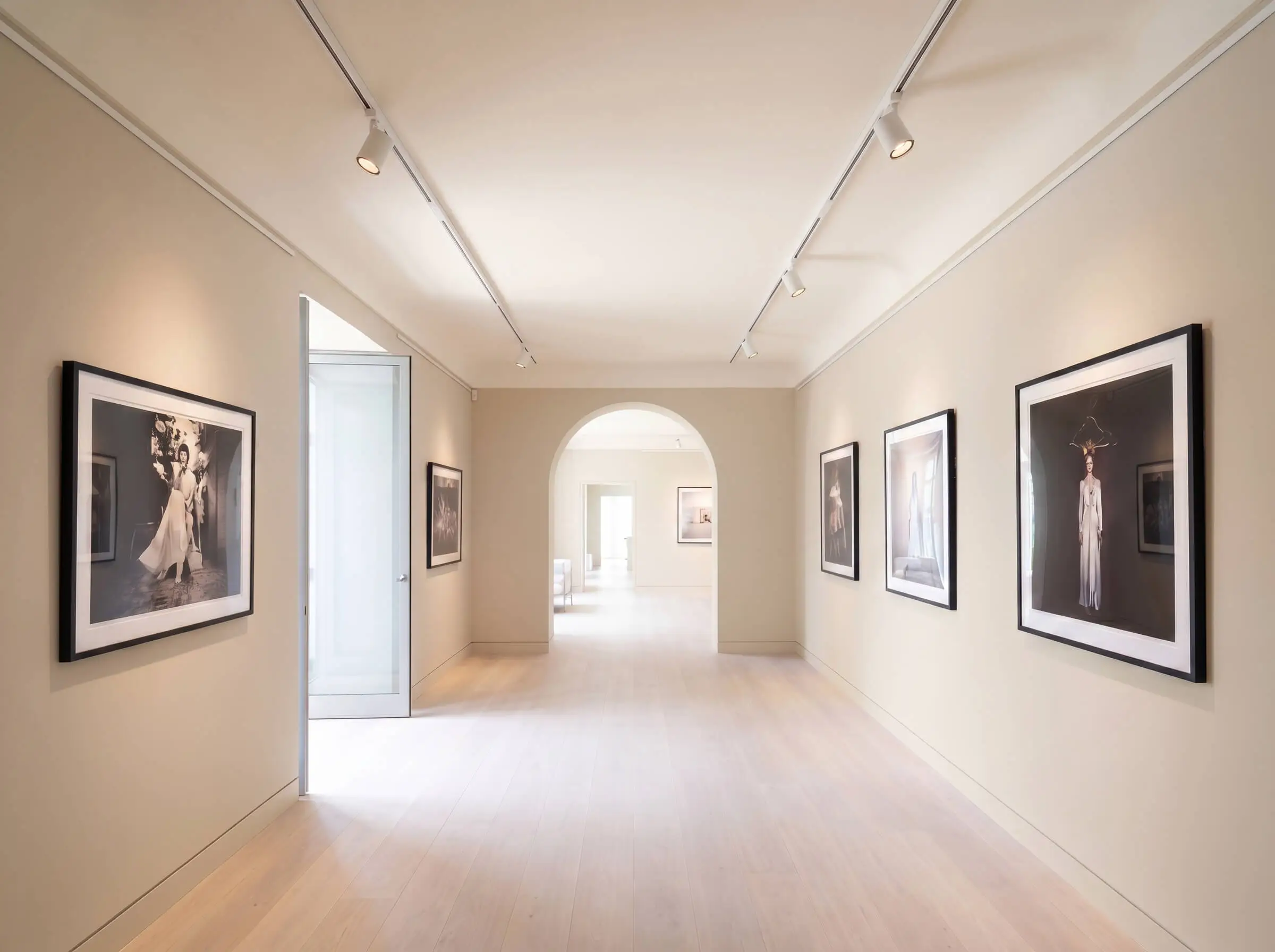 Framed fine art photography lines a minimalist corridor featuring pale beige wall paint, seamless poured concrete flooring, and precise gallery track lighting.