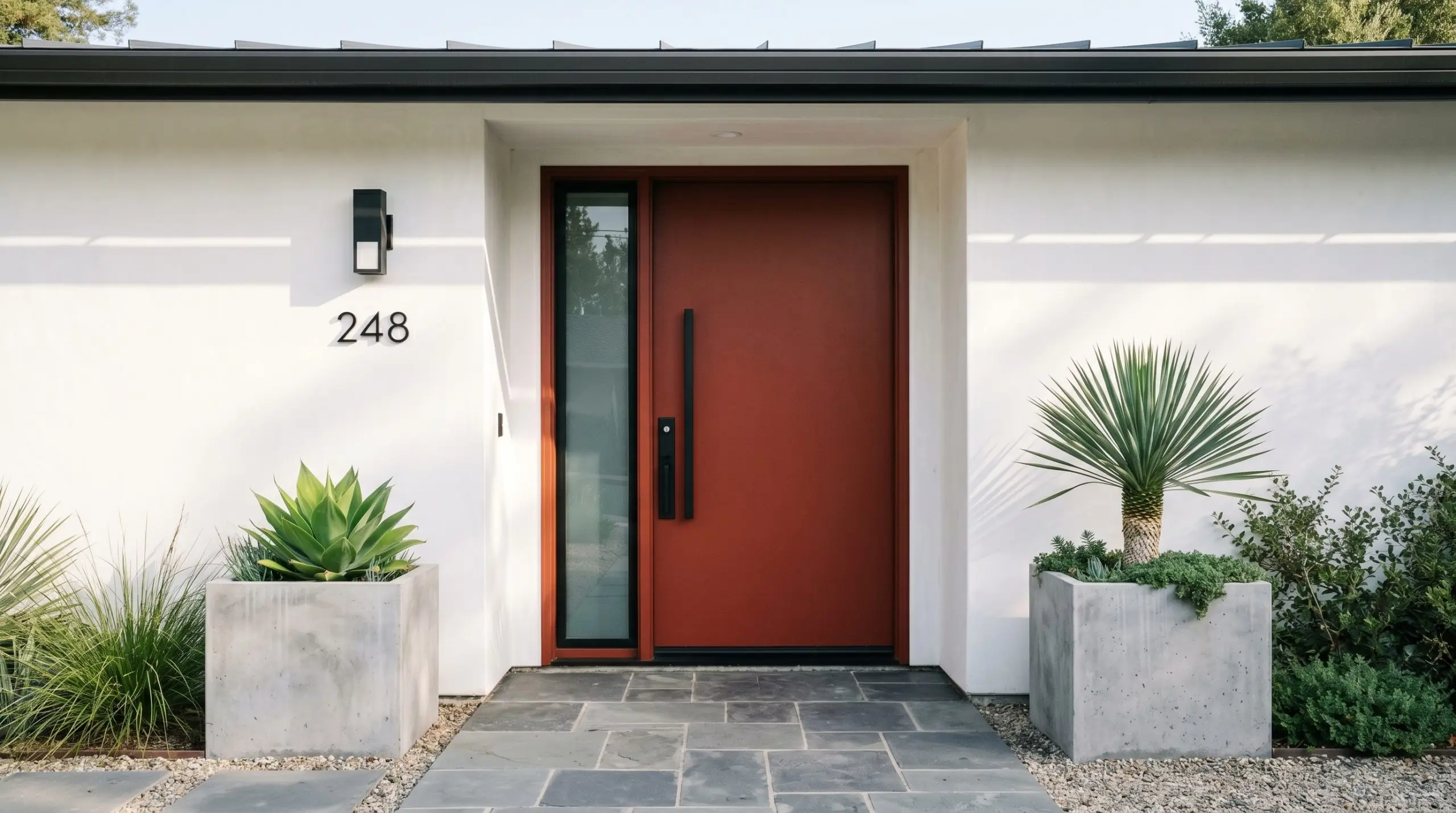 Oversized matte black entry hardware accents a solid brick tone front door set against smooth modern white stucco walls and minimalist concrete planters.