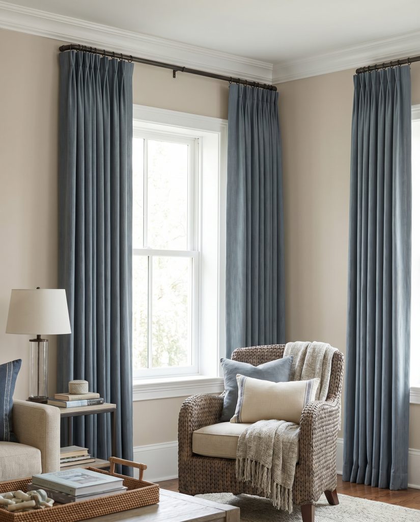 Floor-to-ceiling muted slate blue pinch-pleat drapes hang below crown molding in a sherwin williams accessible beige living room featuring a cozy accent seat.
