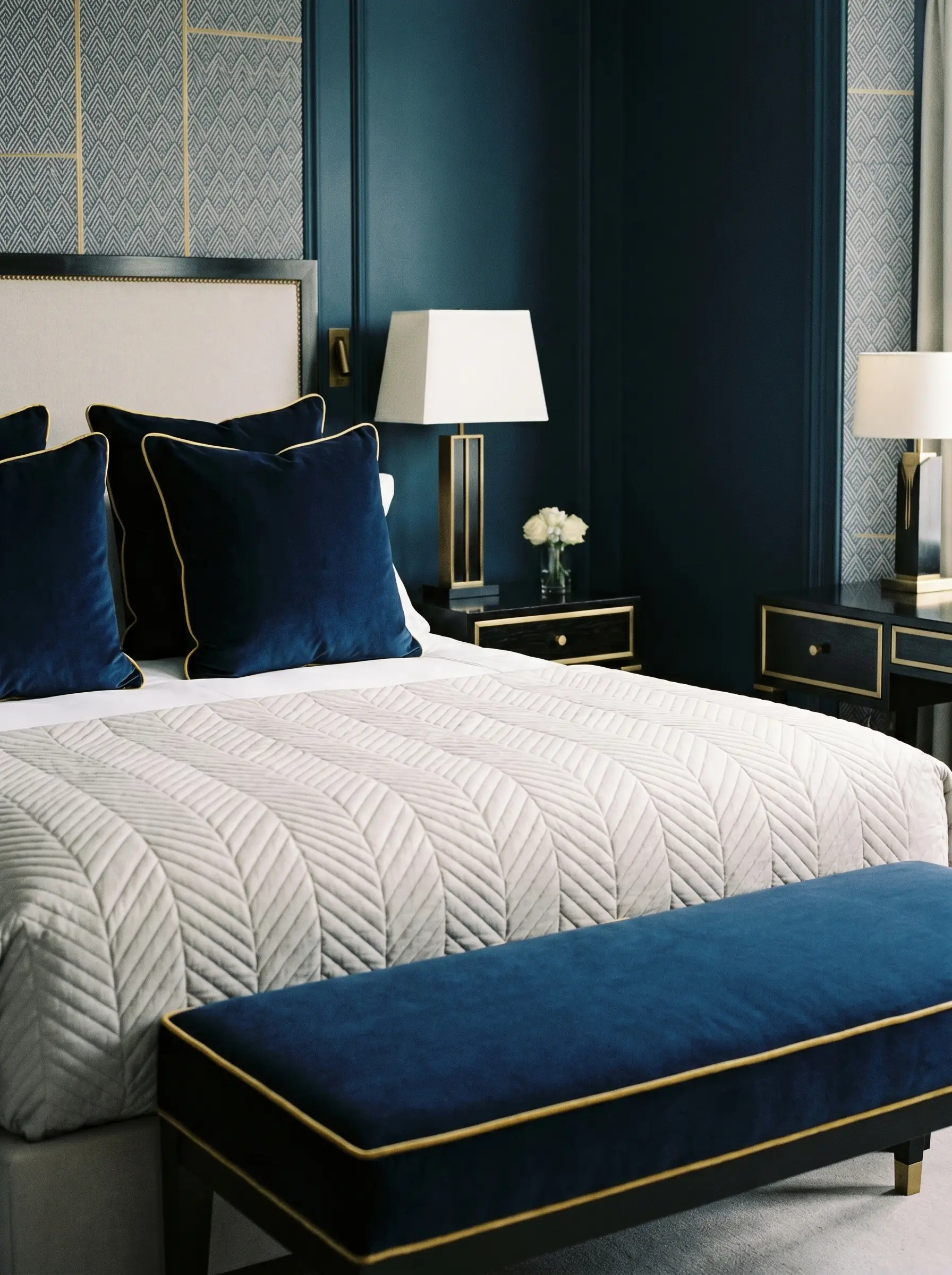 Heavy navy blue velvet pillows with stark gold silk contrast piping rest on a tailored bed behind an upholstered bench featuring matching welt details.