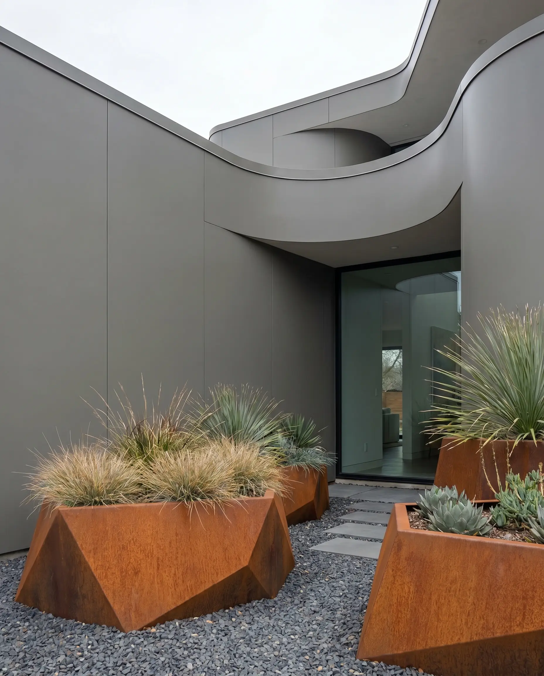 Large, architectural Corten steel planters with a deep oxidized rust patina sit on modern gravel in front of neutral, mid-tone gray siding.