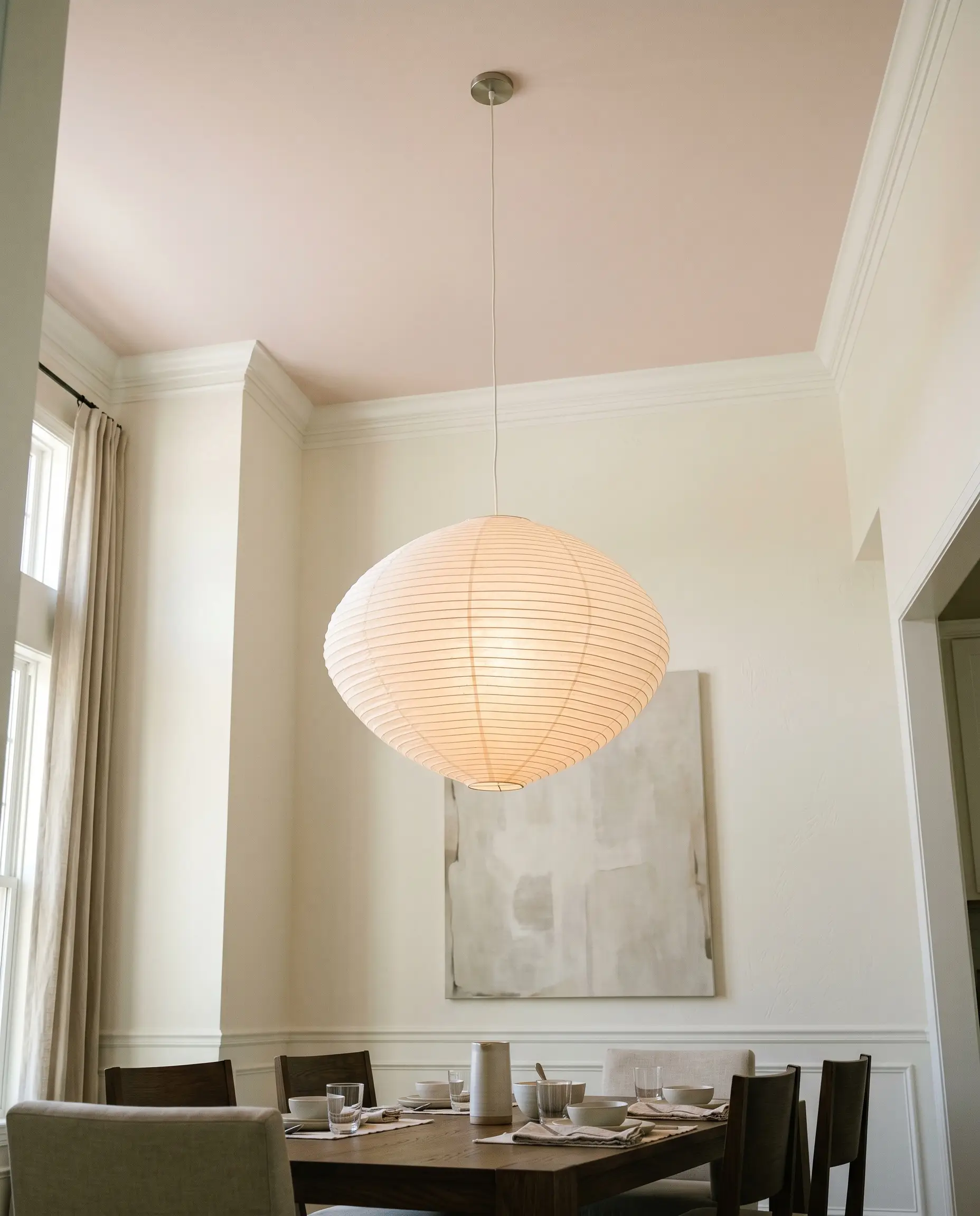 An oversized sculptural paper lantern hangs from a flat ceiling painted in Benjamin Moore First Light (Hex #EFE2DE), contrasting with tall creamy white walls and crown molding in a transitional dining room.