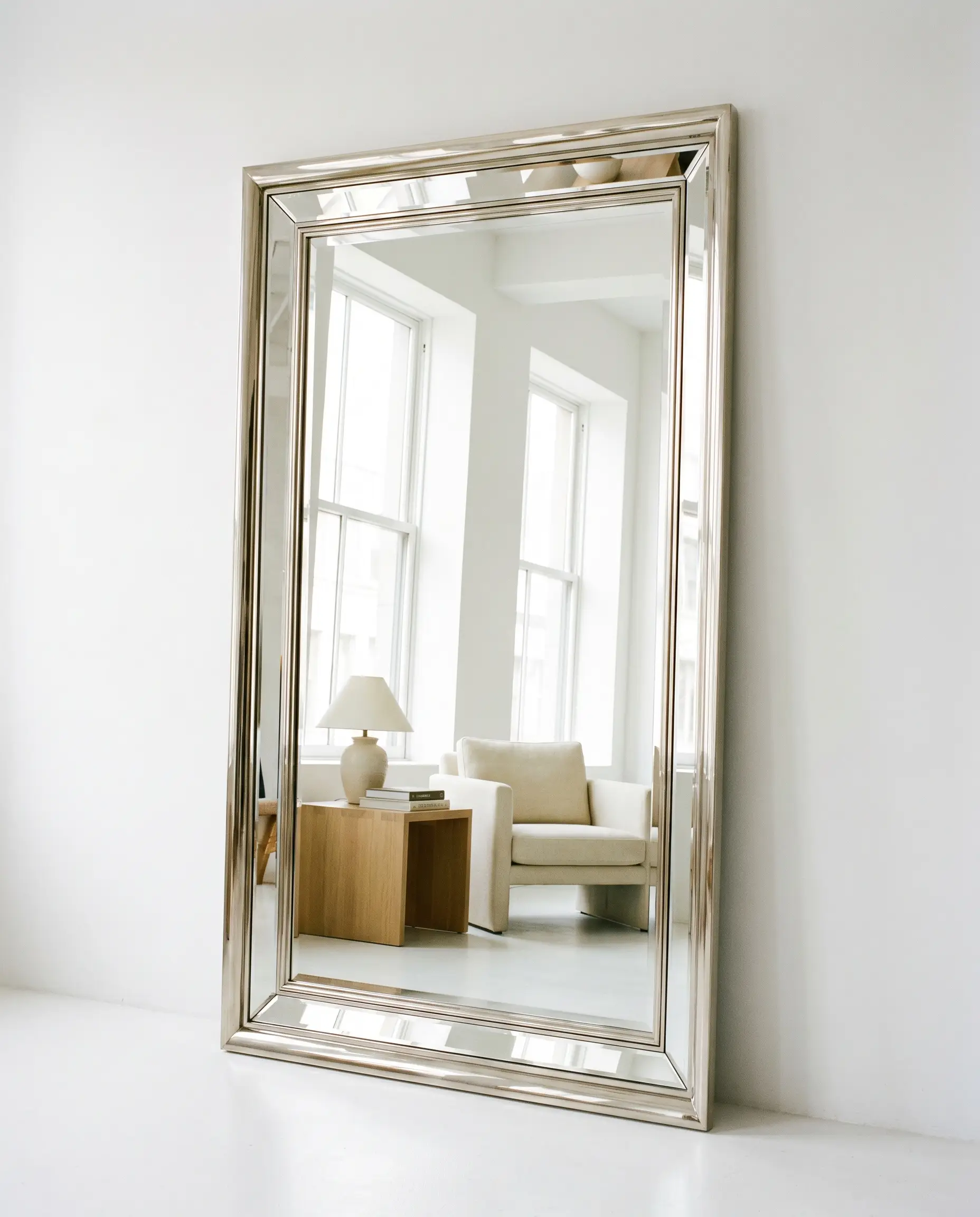 A heavy floor-leaning mirror with a stepped profile and polished nickel border rests against a white wall next to a sleek modern armchair.