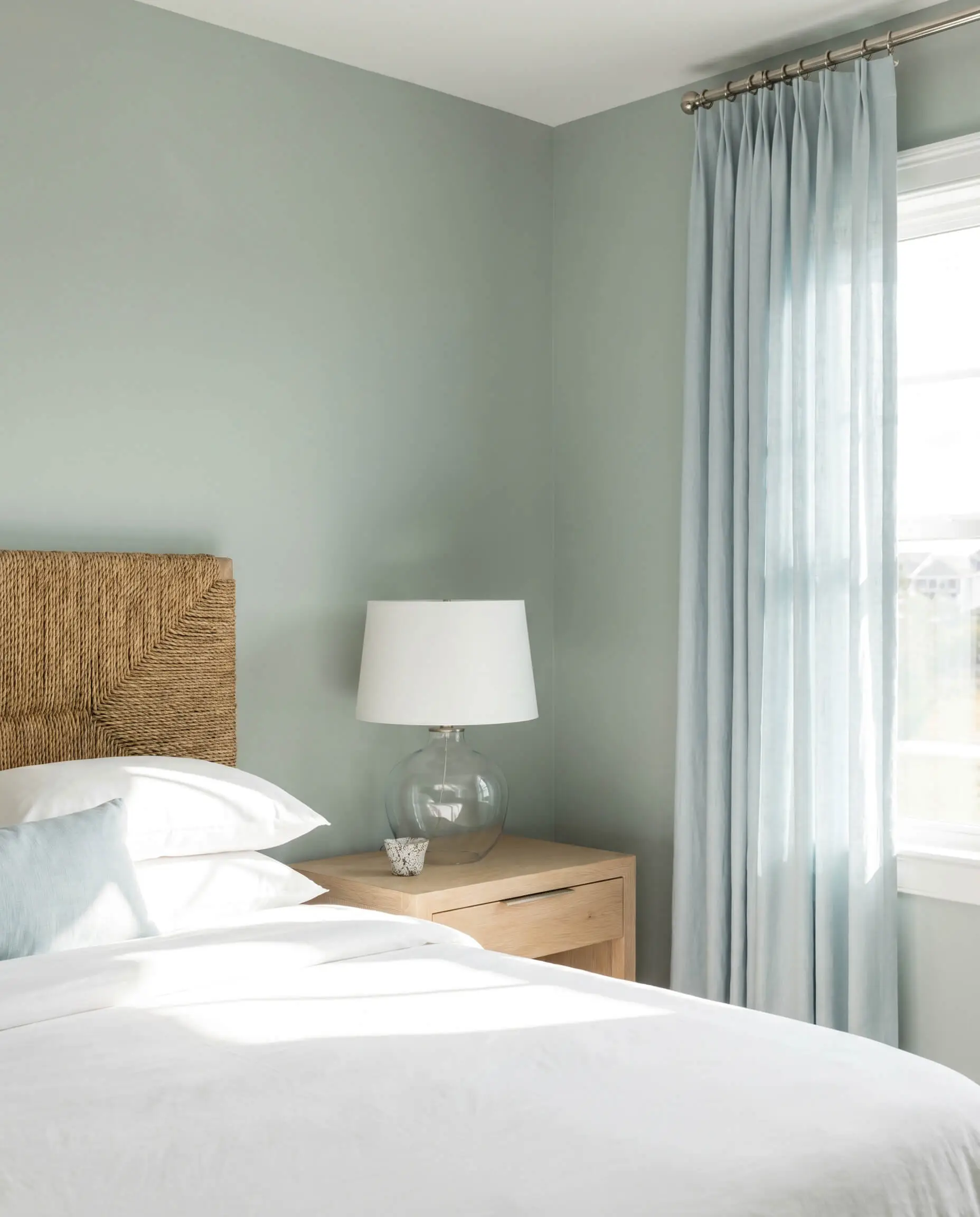 Pale French blue lightweight linen curtains hang from a polished nickel French return rod against Farrow & Ball Cromarty walls near a woven seagrass headboard.