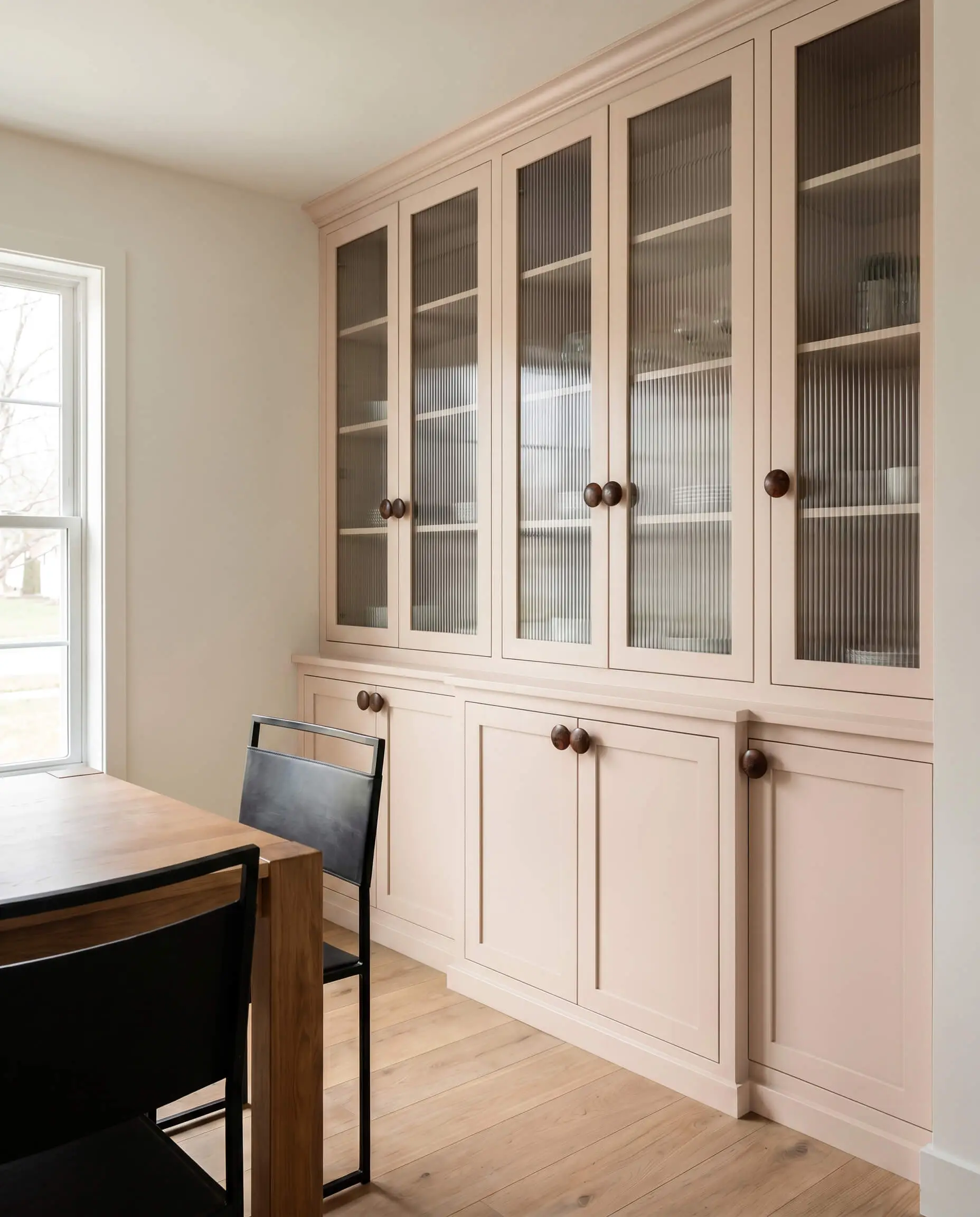 A large built-in dining hutch featuring a sophisticated pastel finish, oversized burl wood knobs, and modern fluted glass doors sits beside a blackened steel chair.