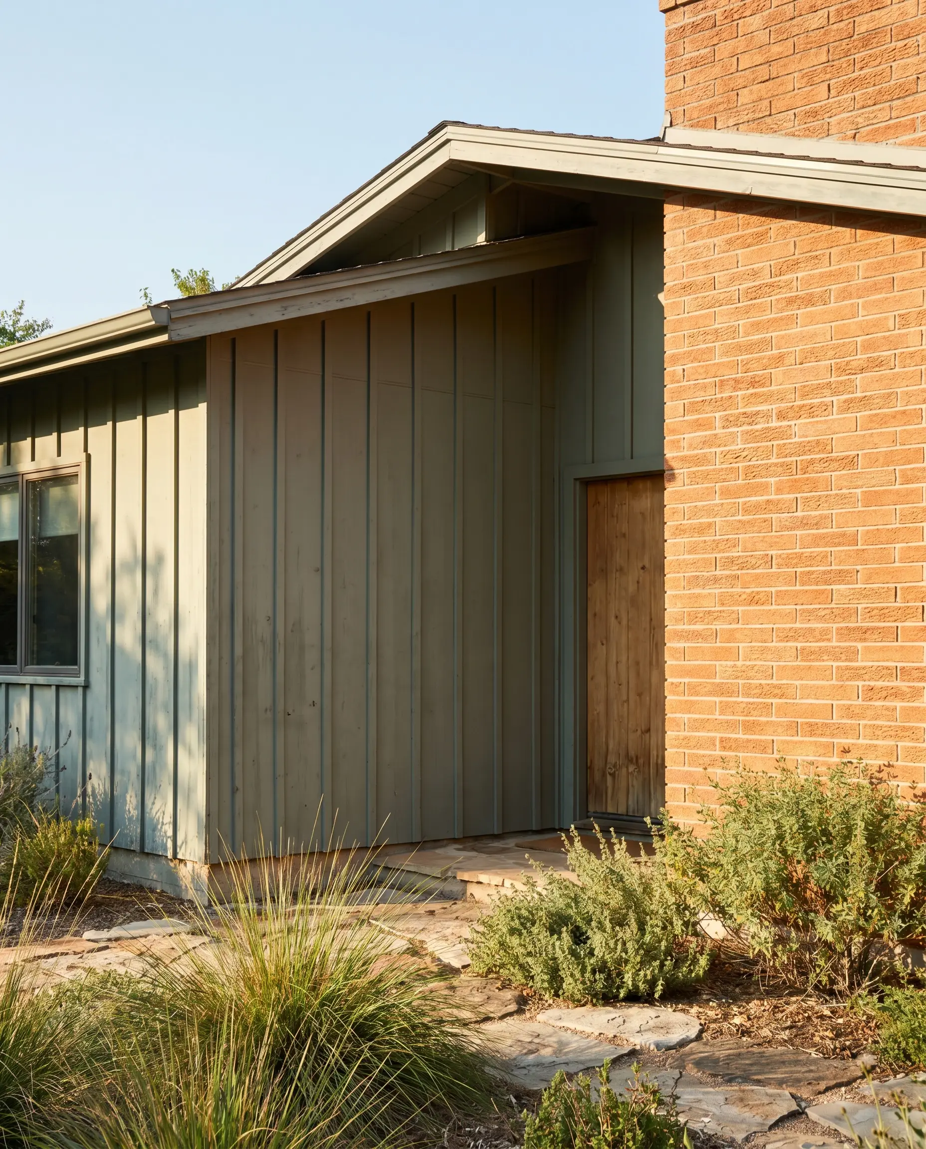 Silvery-green painted board and batten siding meets a fiery orange-red brick exterior wall accented by a simple wooden side door and native landscaping.