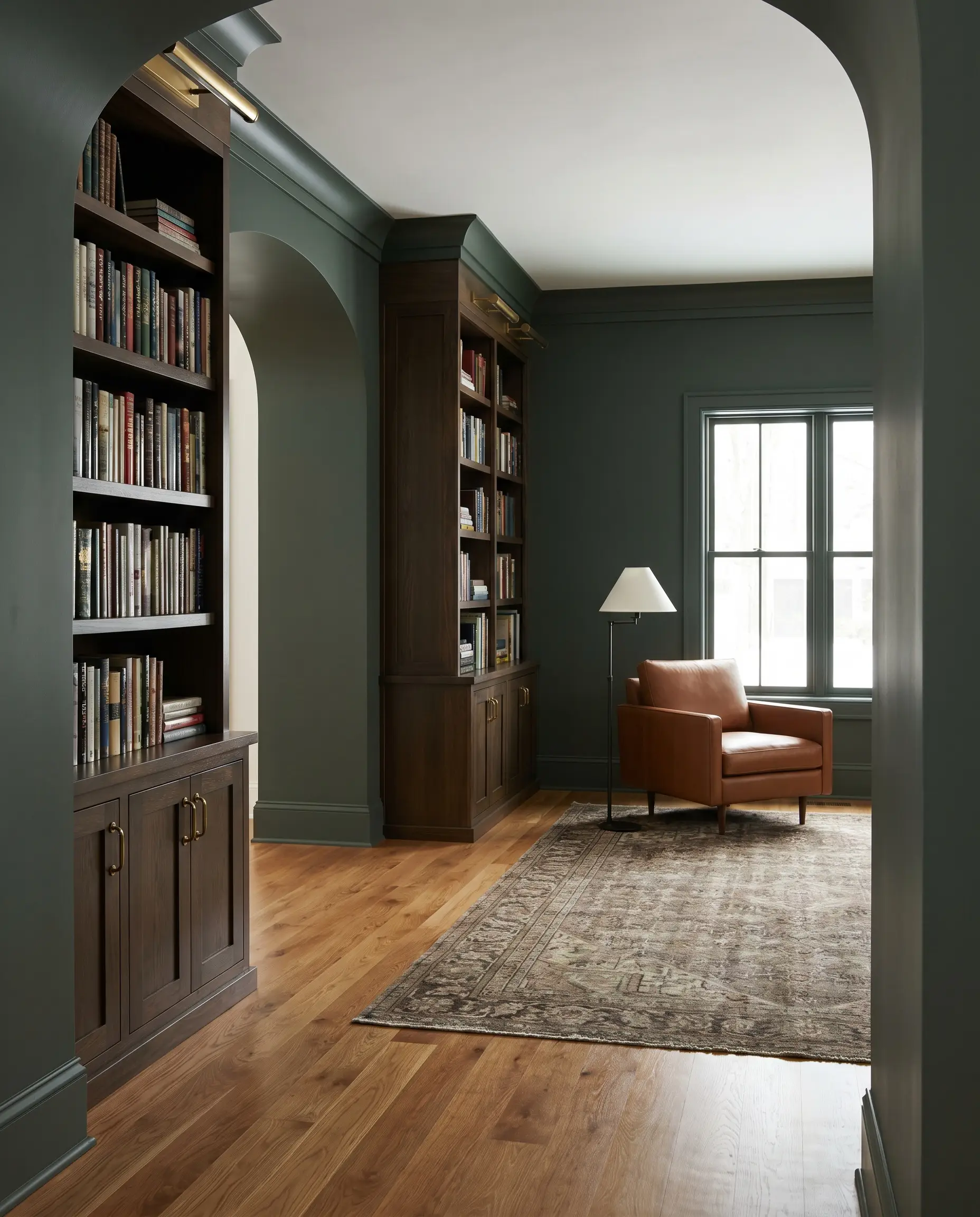 Built-in cabinetry with unlacquered brass hardware and a sleek leather reading chair sit against Sherwin-Williams Pewter Green walls and honey-toned red oak floors.