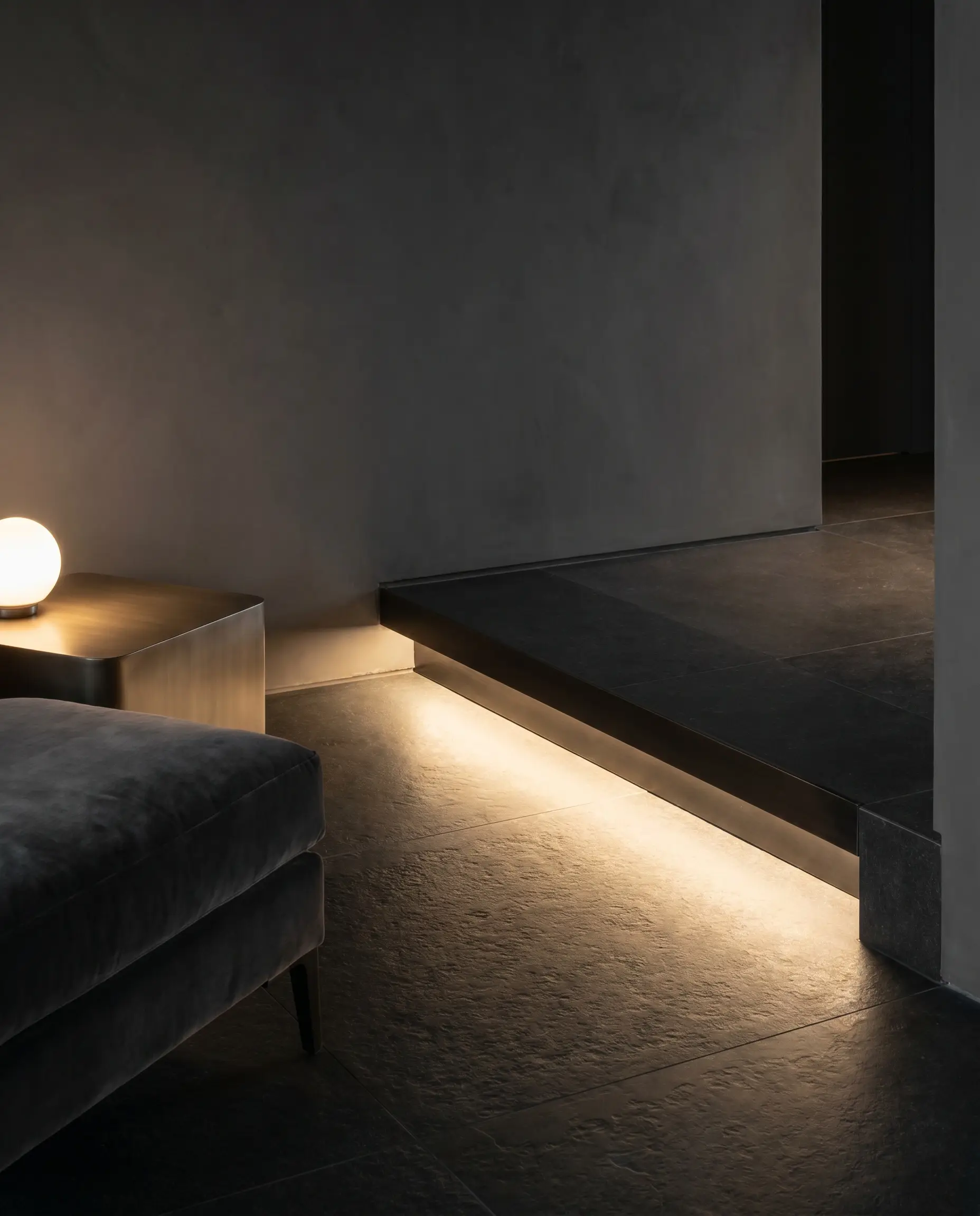 An integrated plaster-in aluminum LED channel illuminates a dark honed slate floor beneath Down Pipe Roman clay walls and a velvet lounge chair.