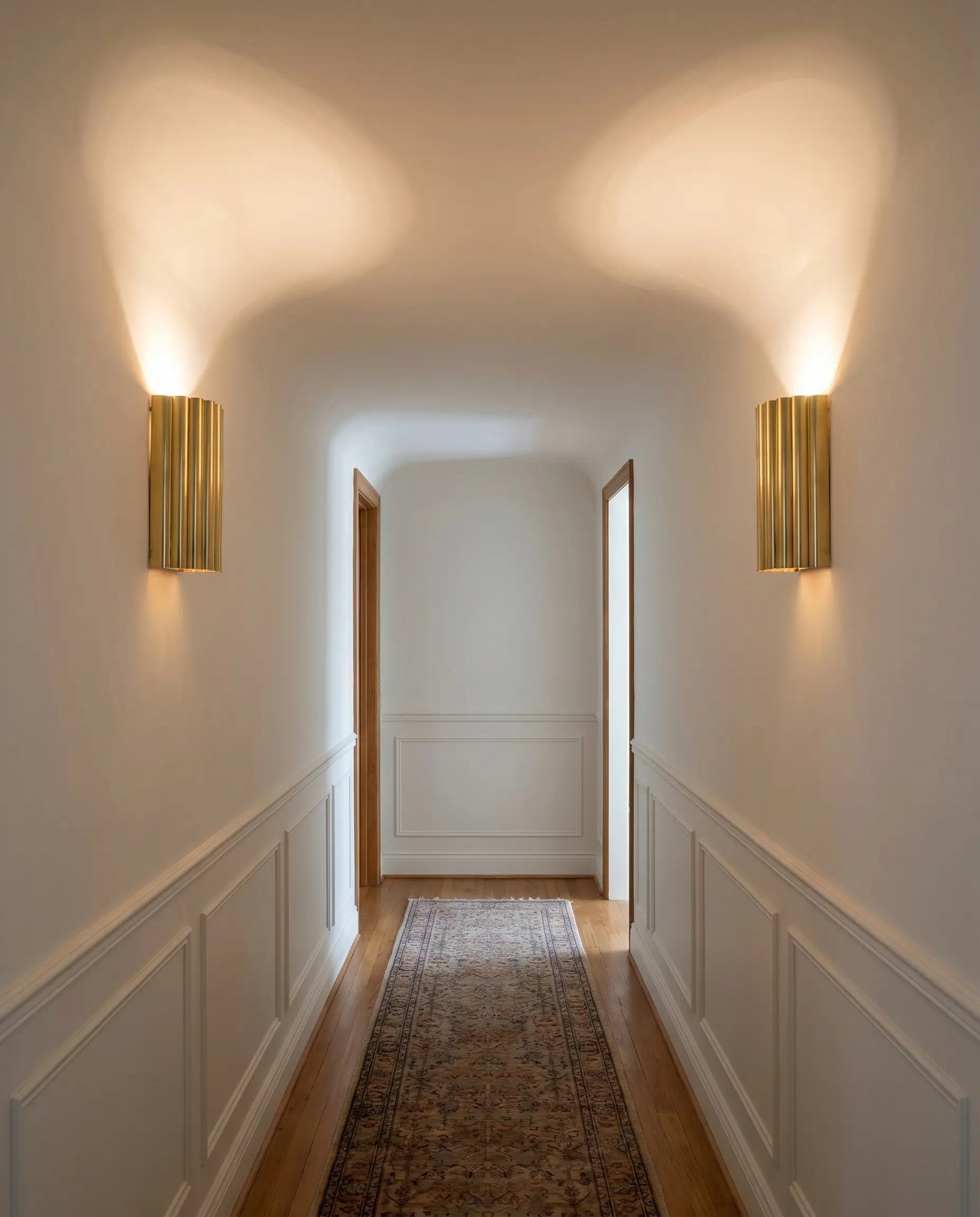 A pair of polished brass fluted half-cylinder wall sconces provides uplighting in a hallway featuring architectural wainscoting and a vintage runner rug.