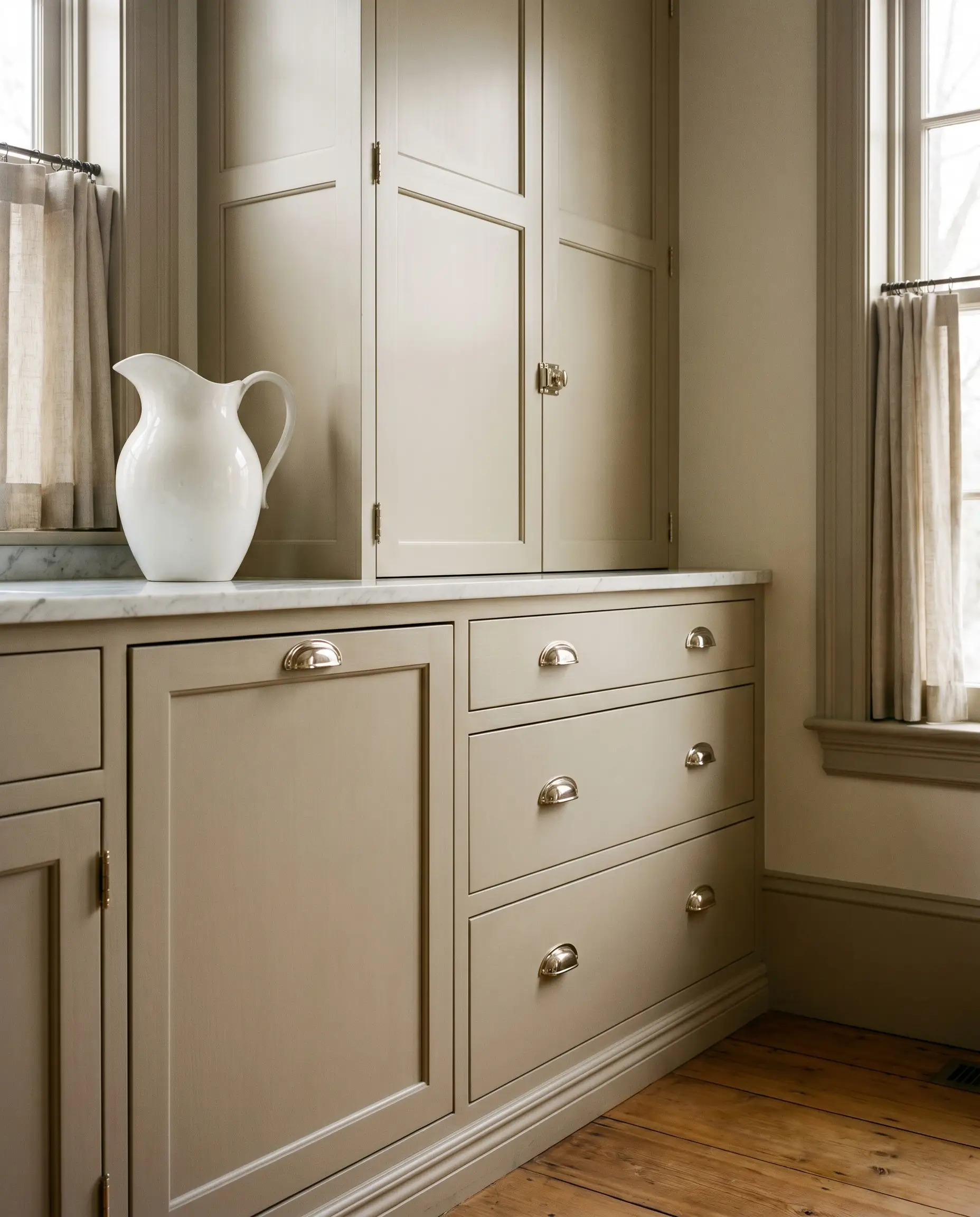 Polished nickel latches, cup pulls, and exposed hinges decorate the millwork of this warm greige kitchen, styled with a classic white ceramic pitcher.