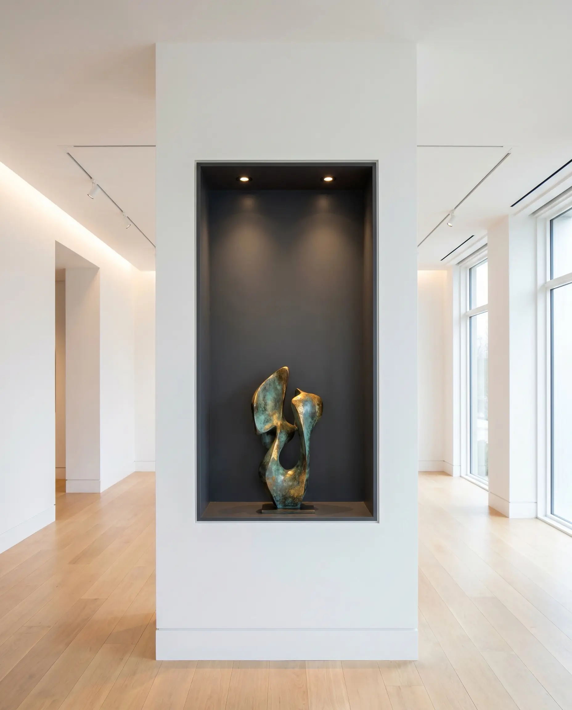 An illuminated recessed display niche built into the drywall cavity of a widened structural support showcases a bronze sculpture in a museum-modern gallery home.