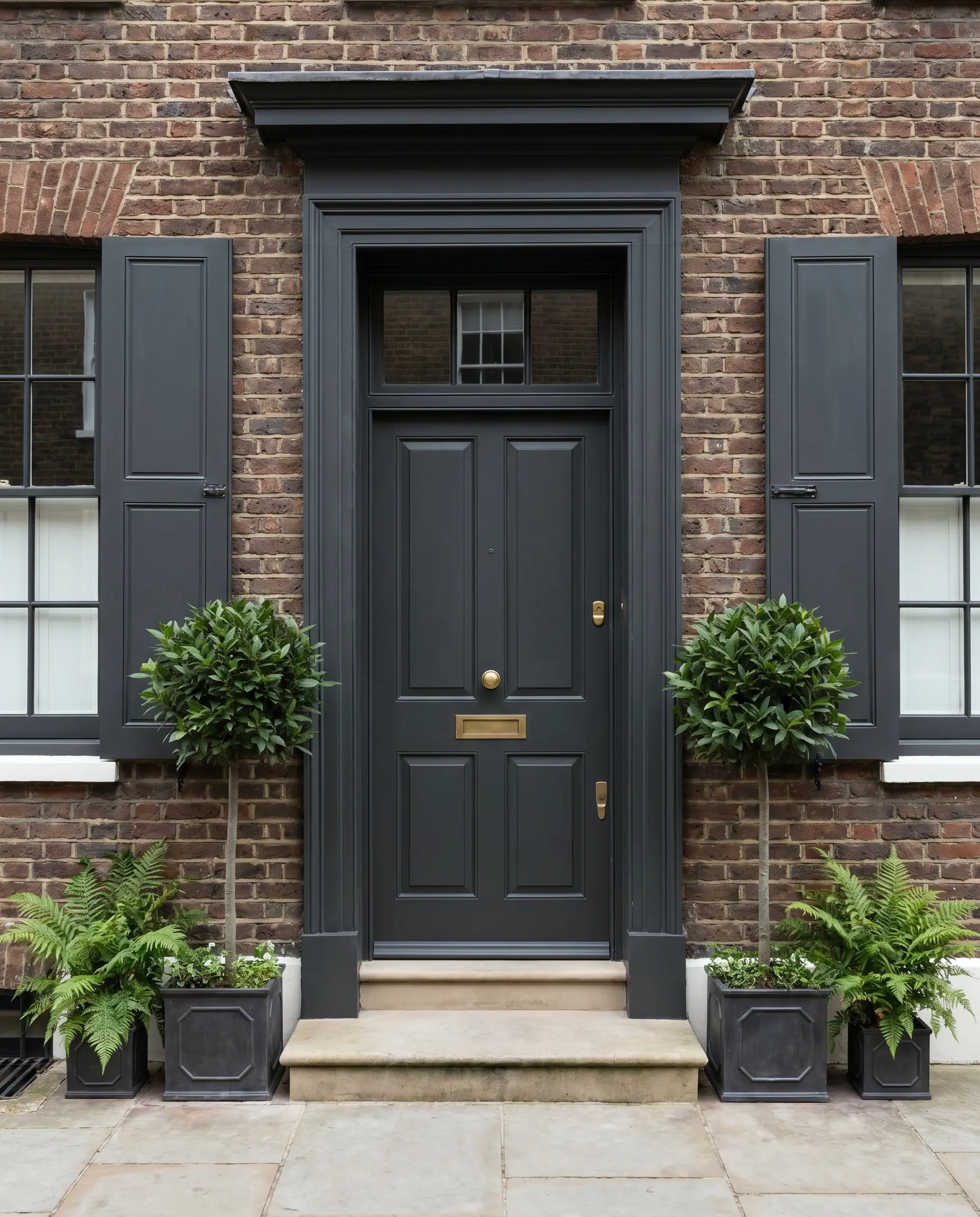 An elegant off-black painted front door and matching operable window shutters contrast perfectly against a rough brown-red brick exterior and brass hardware.