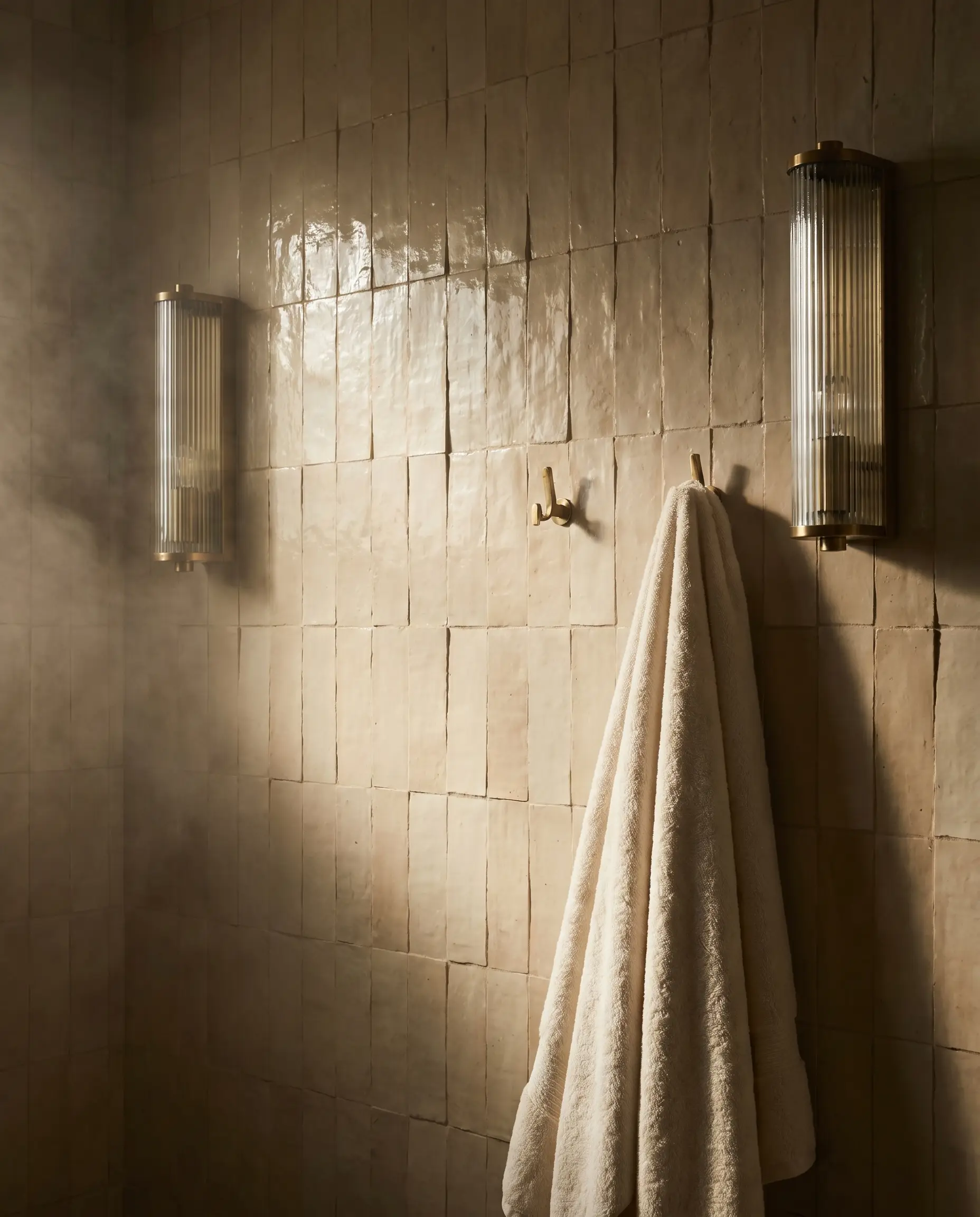 Ribbed glass and brass wall sconces mount vertically on rectangular clay tiles, illuminating the natural lippage and unrectified edges of the artisanal surface.
