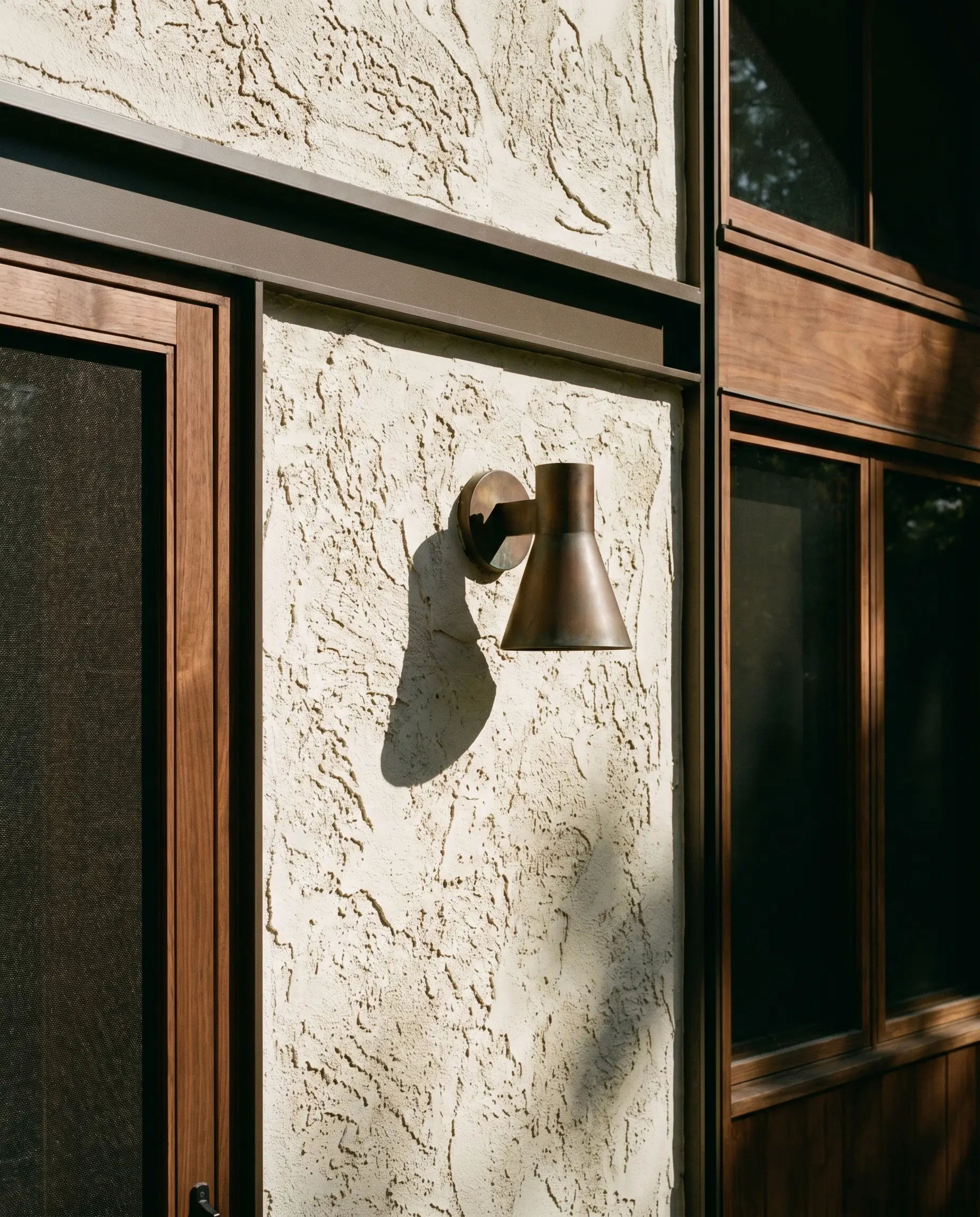 Rough cream stucco rendering contrasts against dark bronze metal trim, solid walnut wood elements, and a subtle bronze outdoor sconce.