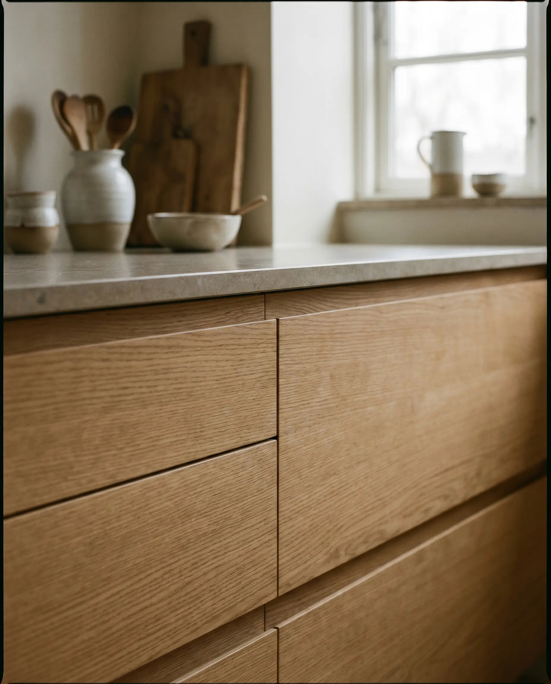 Handleless warm ash wood kitchen cabinetry features integrated J-channel routed finger pulls that maintain an unbroken horizontal wood grain above a matte stone countertop.