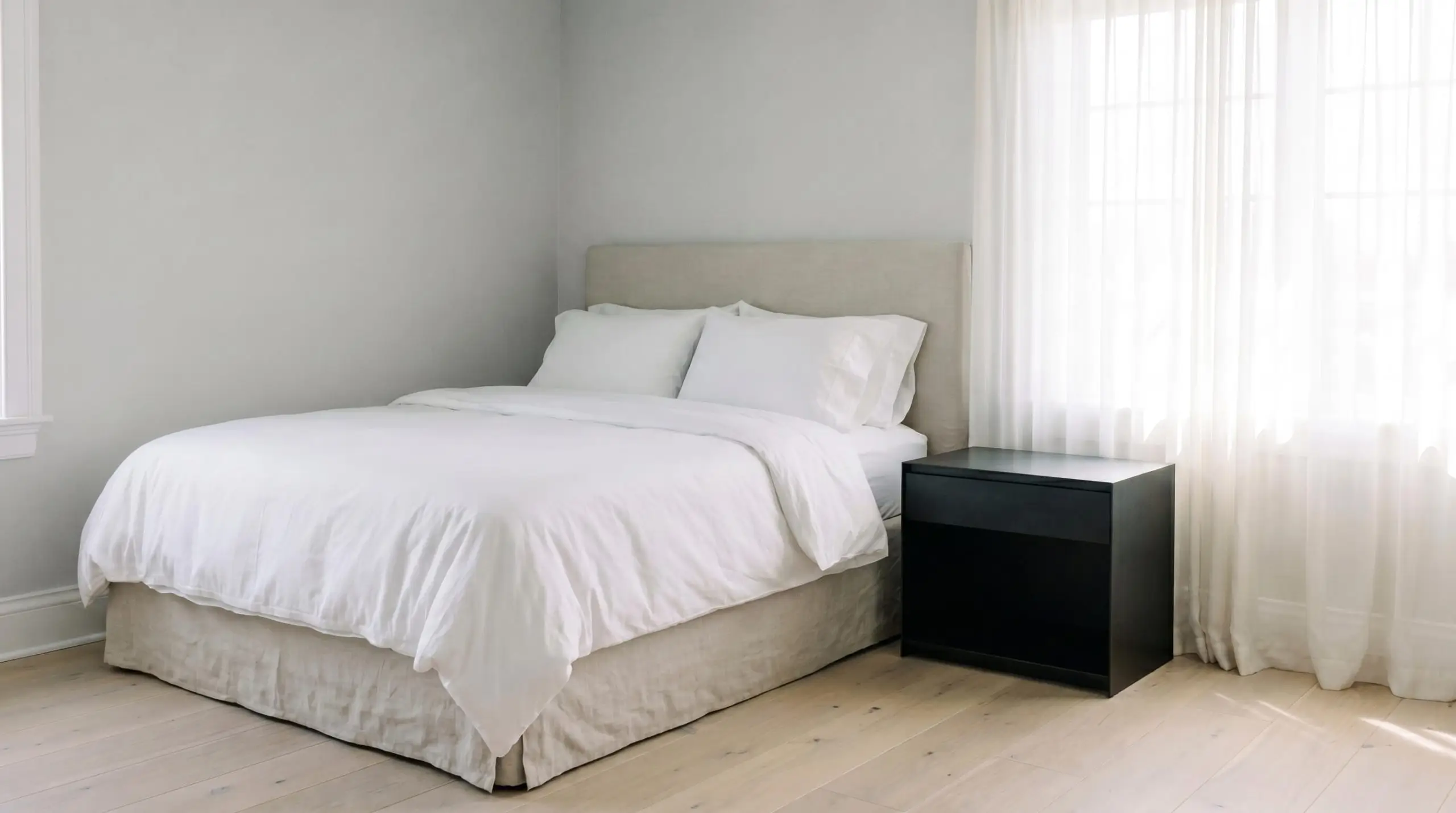 A slipcovered linen bed dressed in crisp white sheets pairs with a blackened steel nightstand against a steely cast architectural finish.