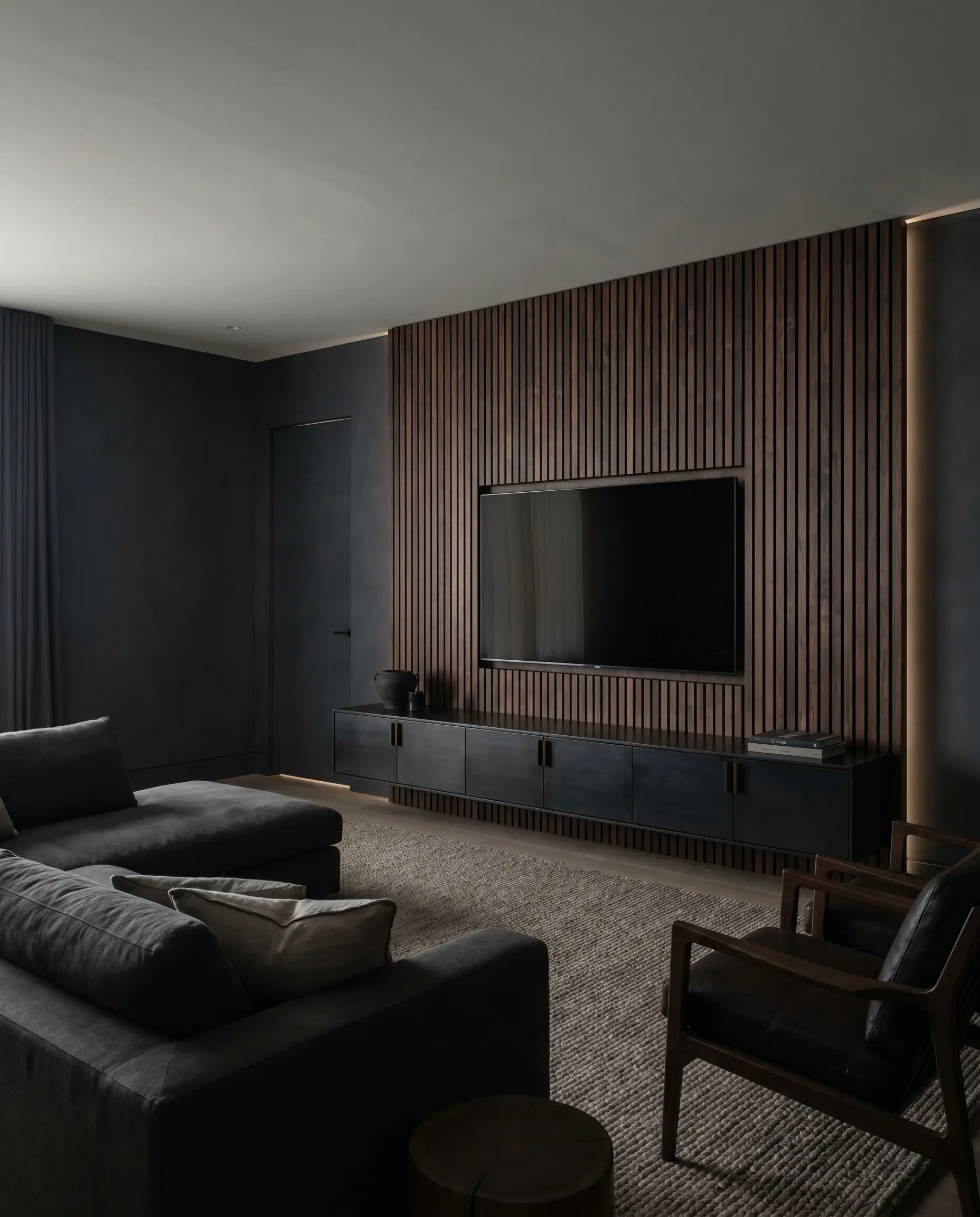 Flat-cut smoked walnut veneer slats back a wall-mounted television and a floating media console in a moody Japandi style wood slat TV walls setting.