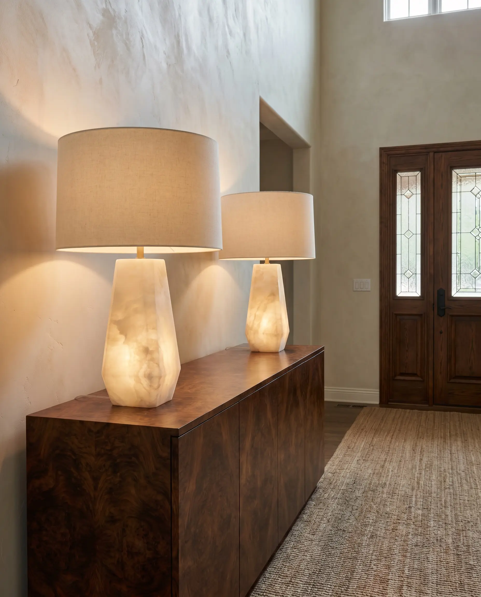 Oversized solid carved alabaster geometric table lamps with linen drum shades rest elegantly on a dark, swirling burl wood console table.